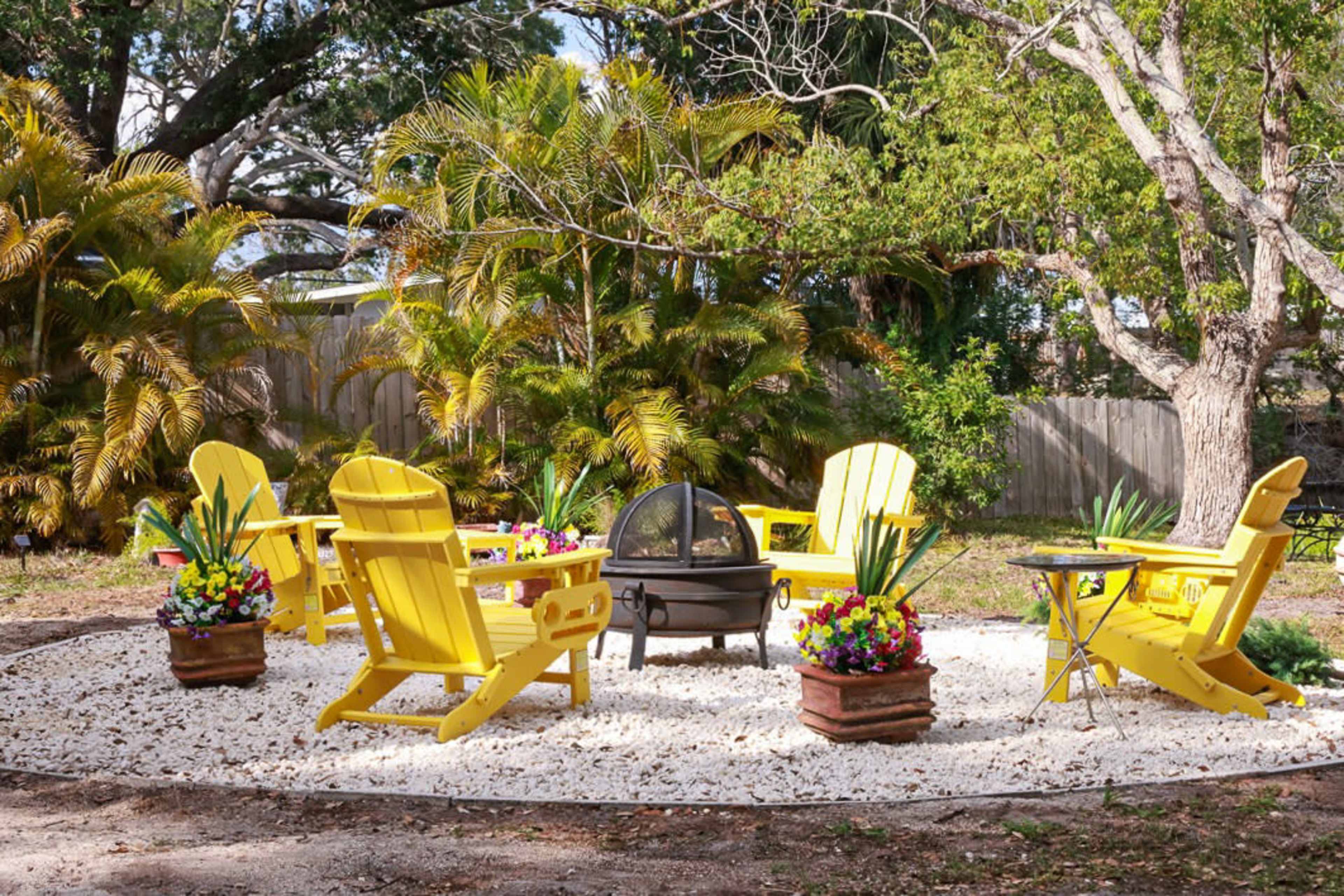 The image shows a circular seating area with five yellow Adirondack chairs arranged around a fire pit, surrounded by potted plants and gravel.