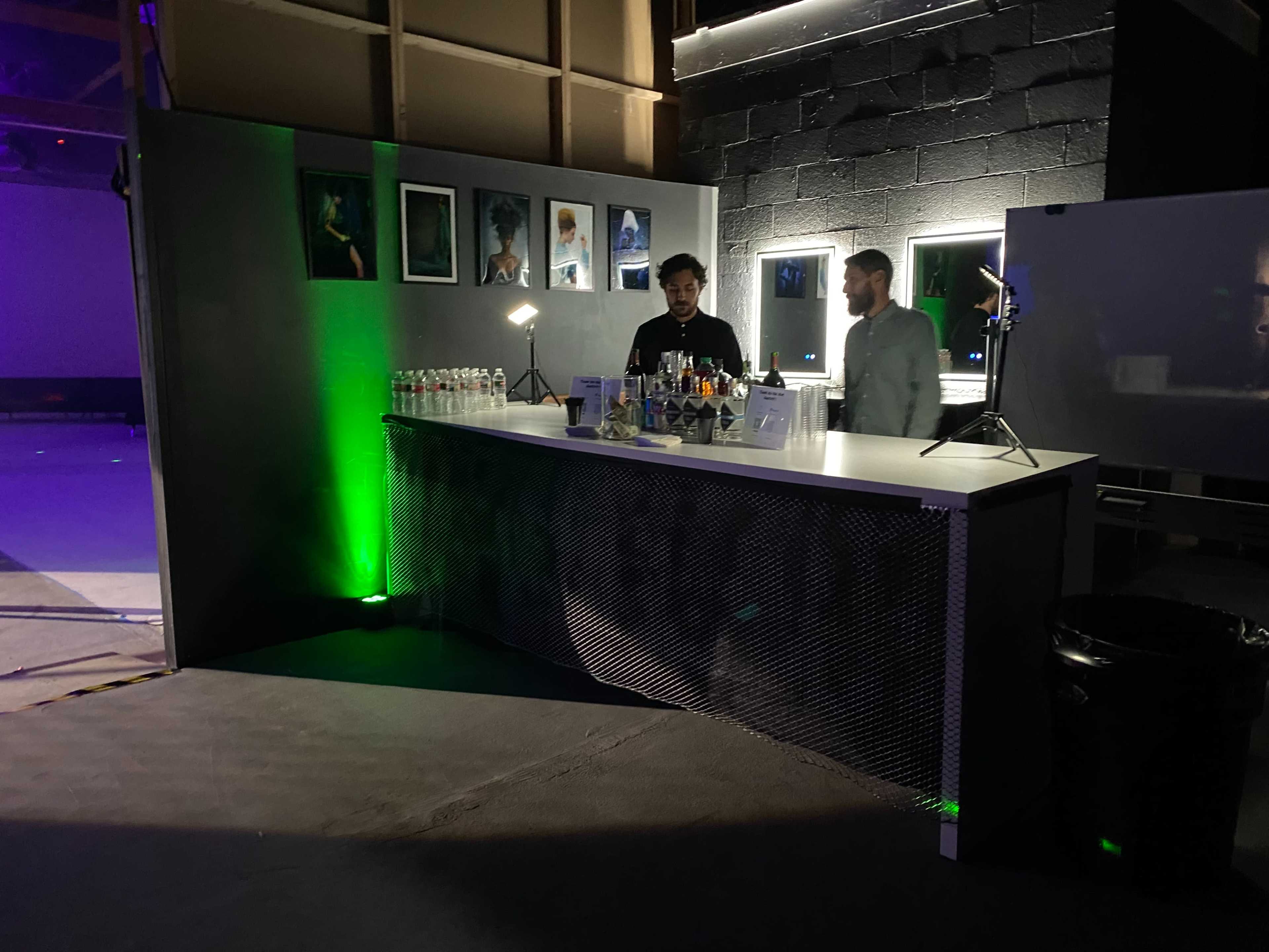 A bar setup is shown with two individuals behind it, surrounded by bottles and beverages, in a dimly lit venue featuring framed artwork on the walls.