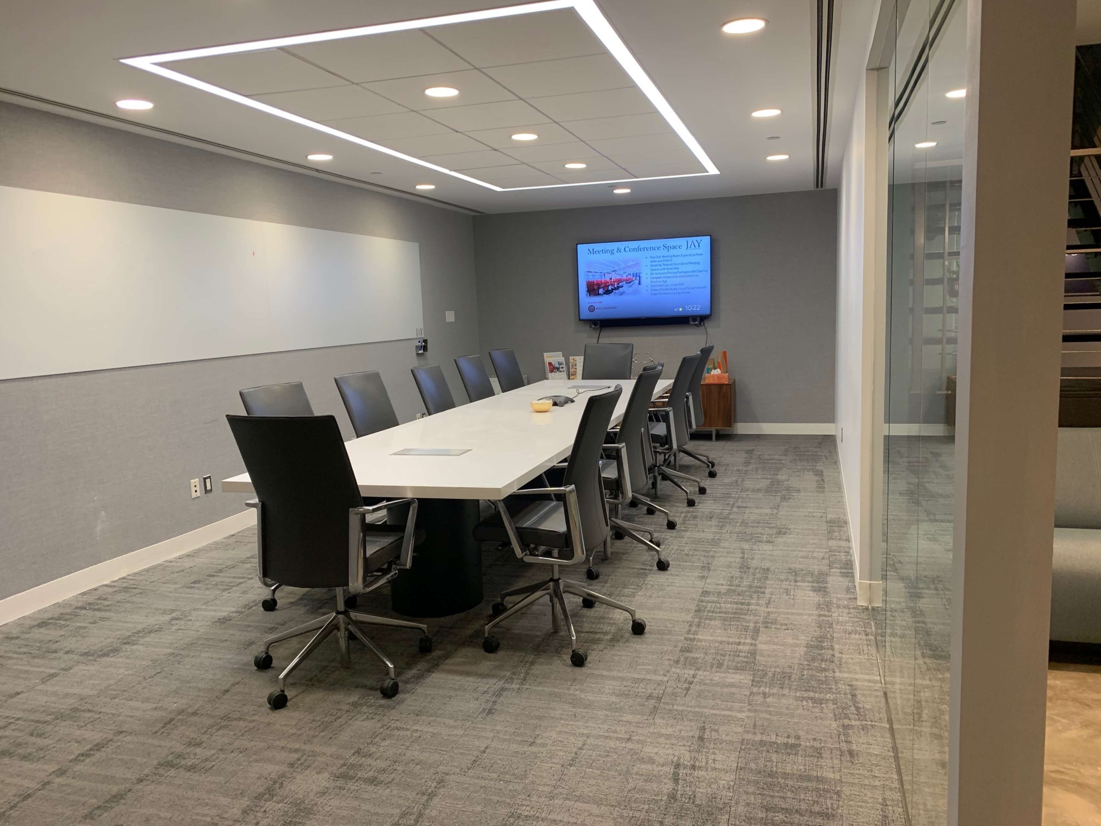 A modern conference room features a long table surrounded by black office chairs, with a screen displaying a presentation on the wall.