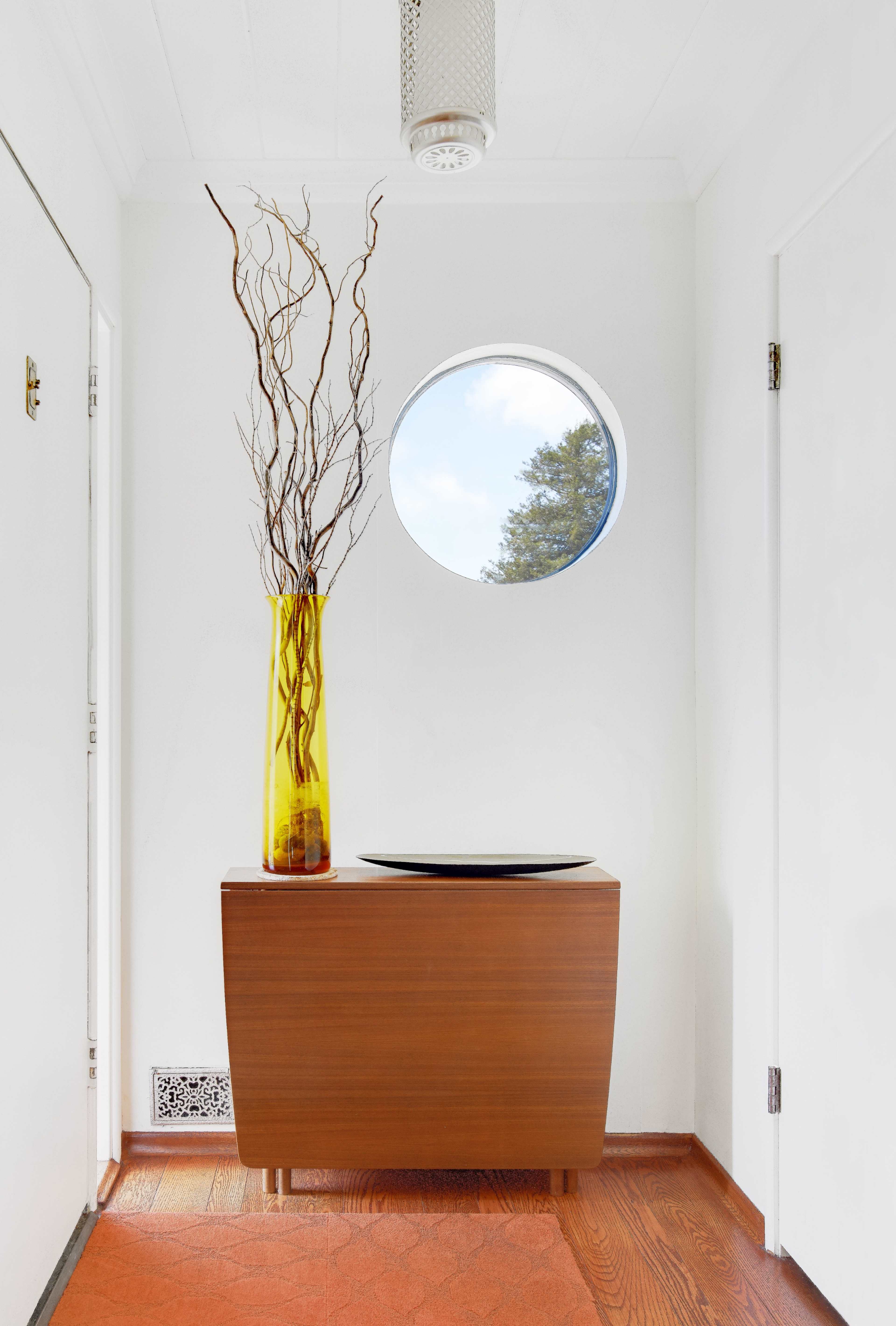 A minimalistic hallway featuring a wooden console table with a round vase containing branches and a circular window illuminating the space.