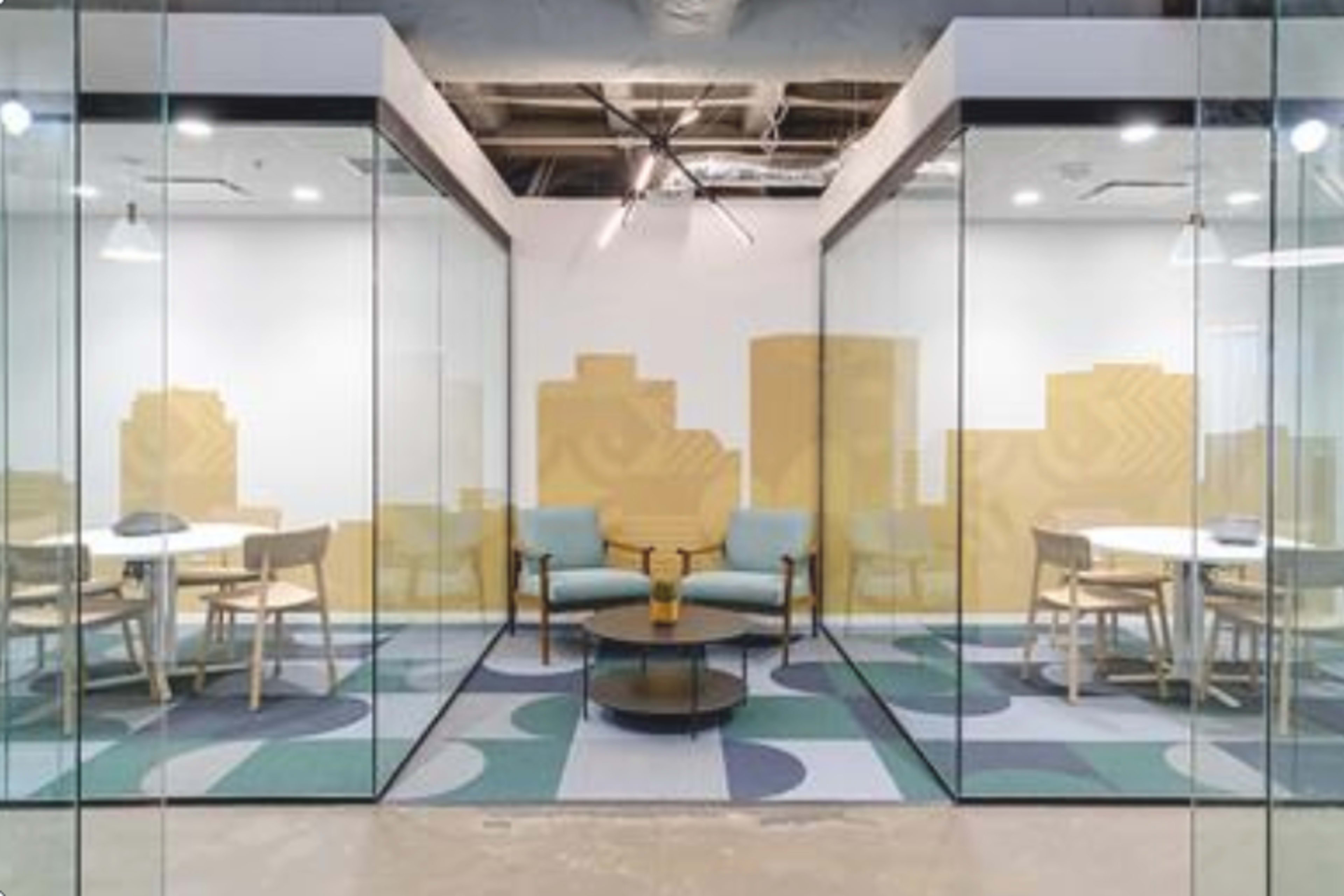 The image shows a modern office space with glass-walled meeting rooms, featuring round tables and light blue chairs on a patterned floor.