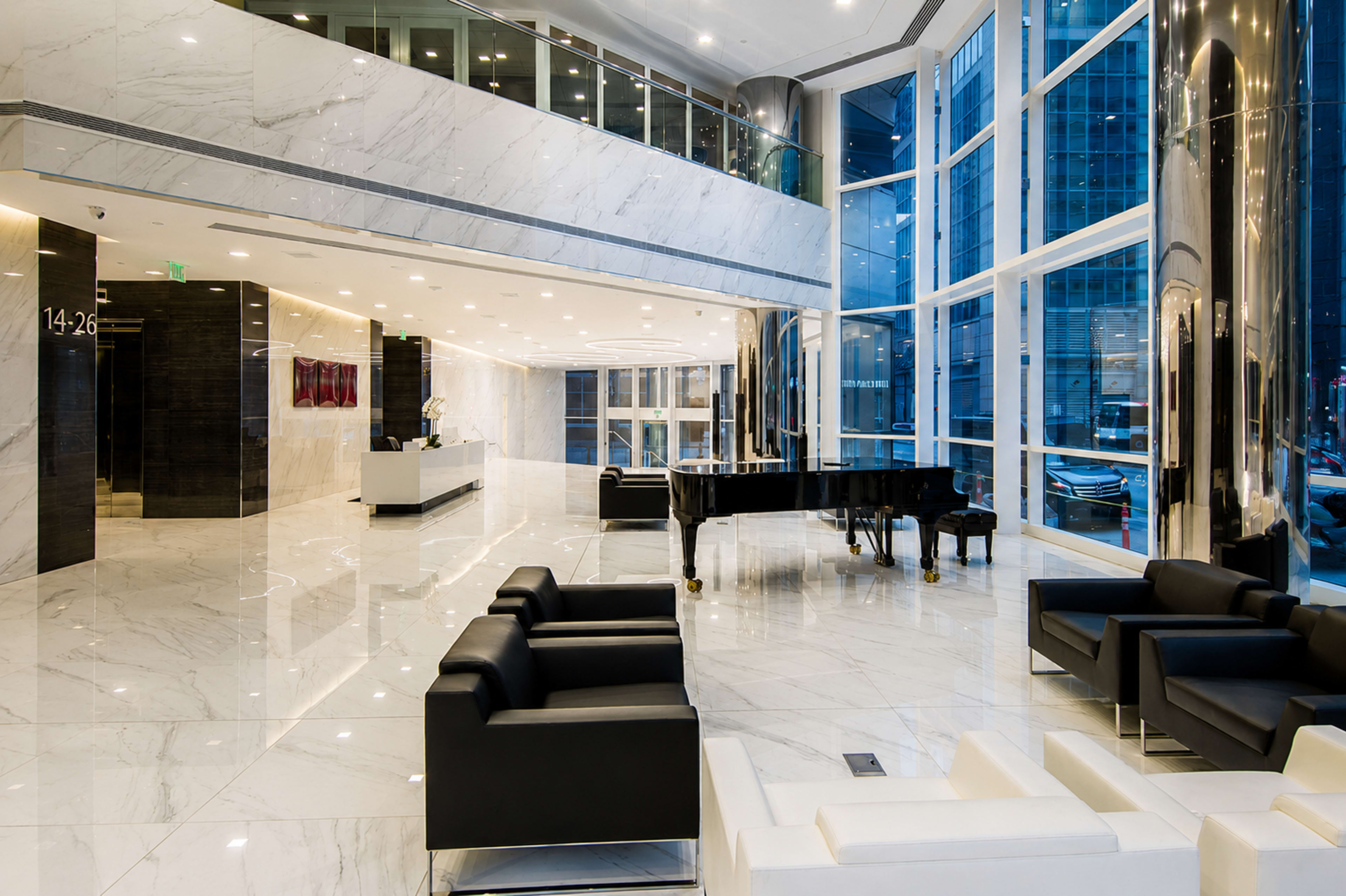 The image shows a modern lobby with marble floors, large windows, sleek black seating, and a grand piano.