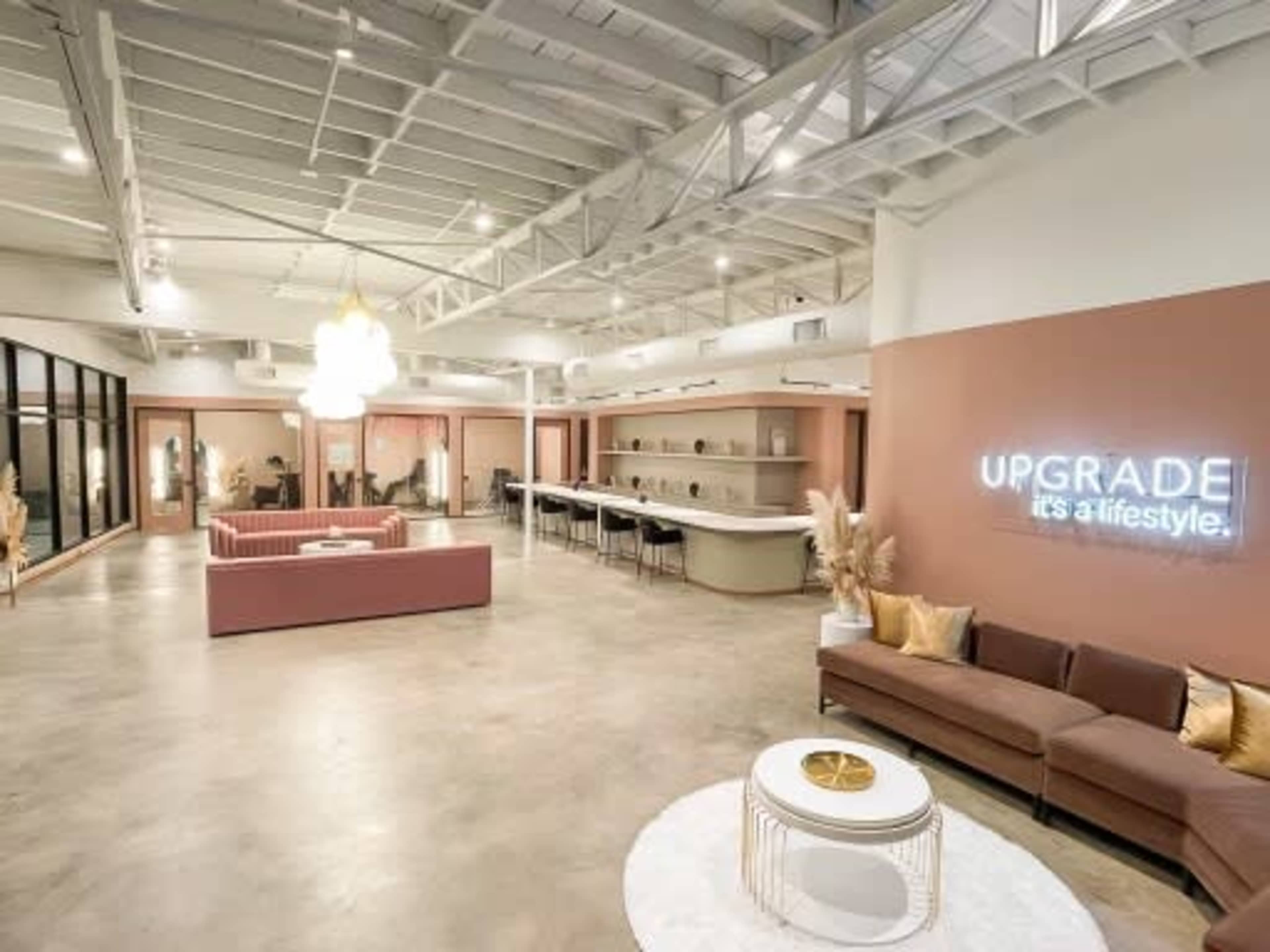 A spacious, modern interior with a pink and beige color scheme, featuring a lounge area with a sofa, a large bar counter, and decorative elements like a neon sign and plants.