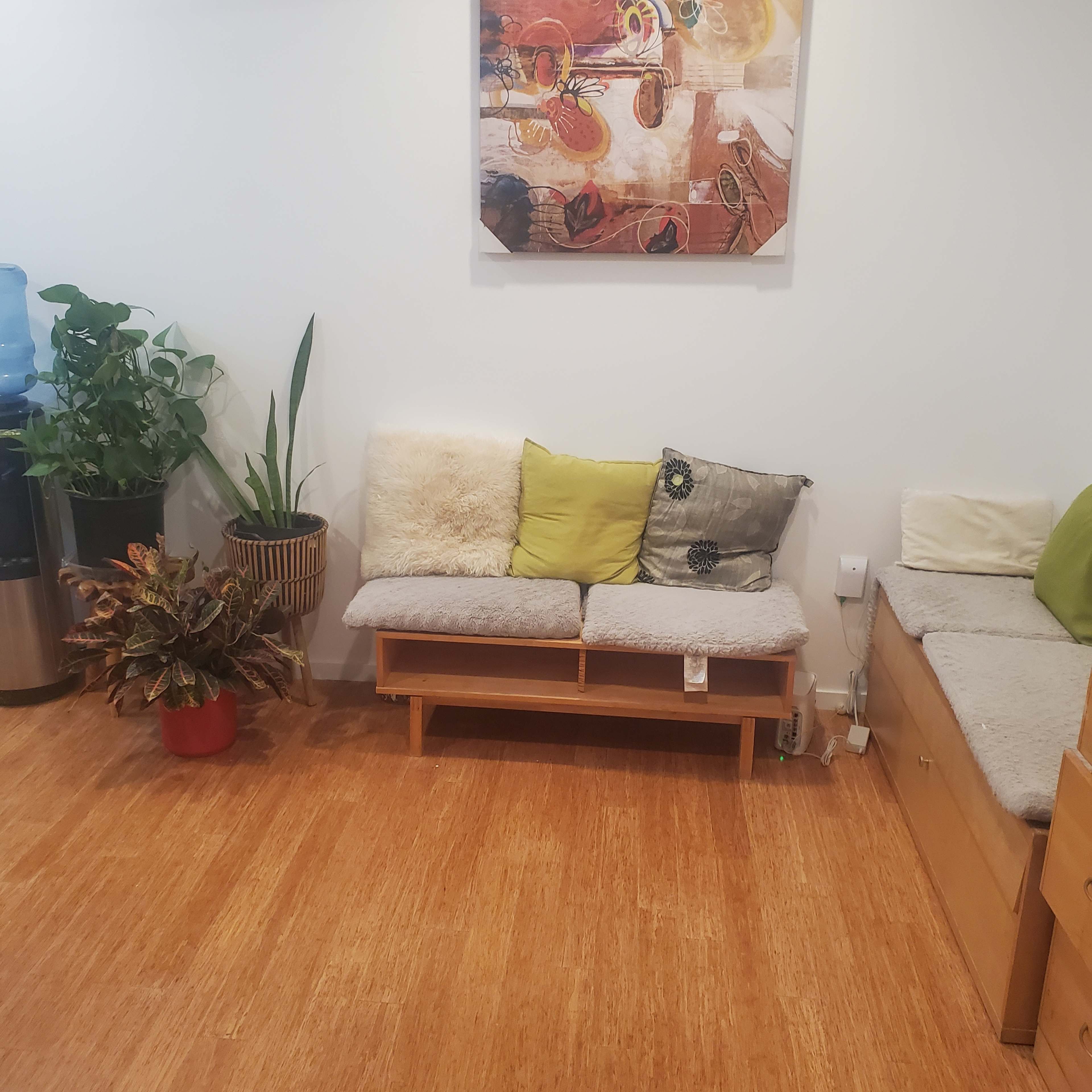 The image shows a room with two wooden benches covered with cushions, a small table, and potted plants, alongside a water cooler against one wall.