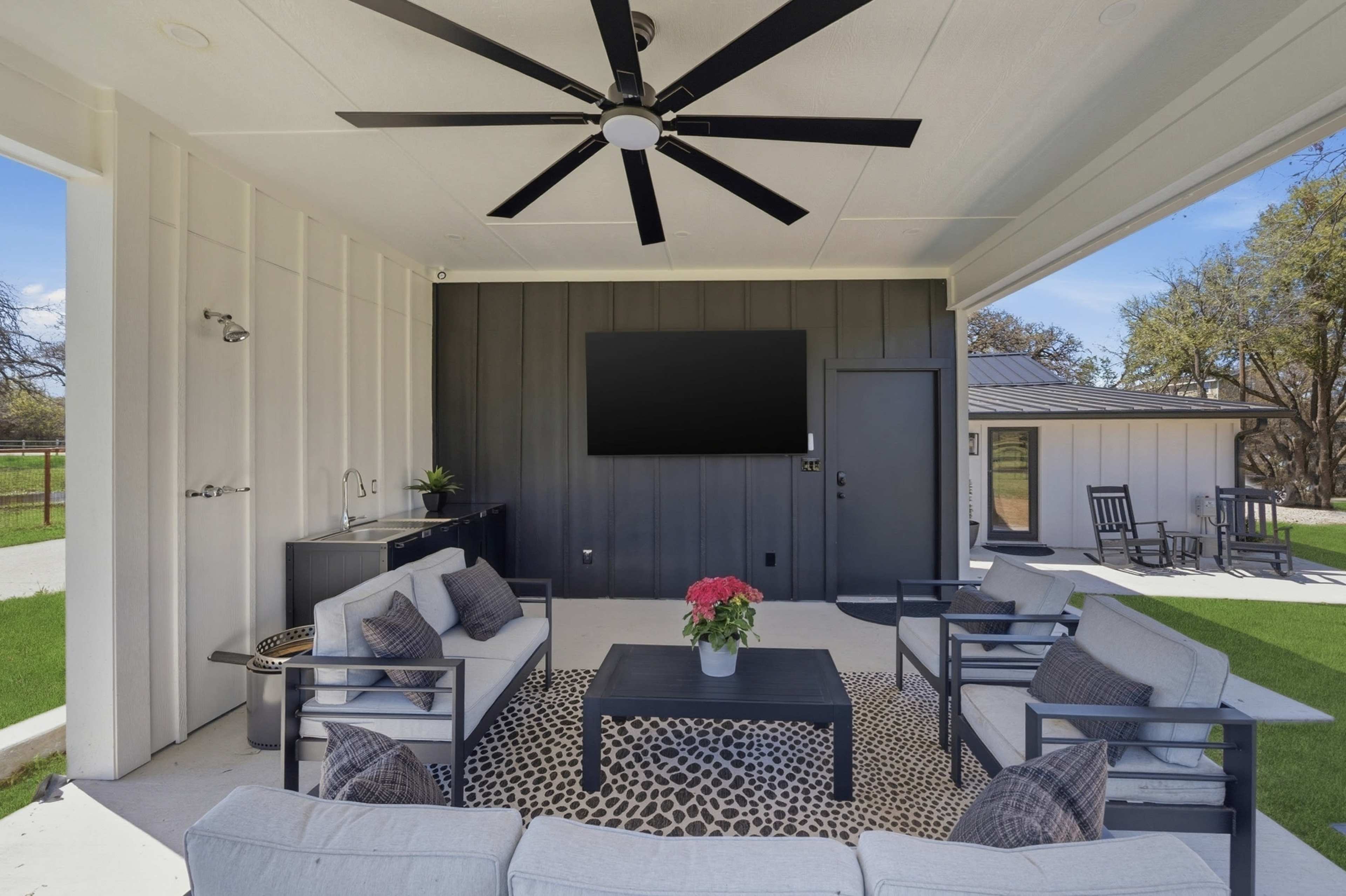 The image shows a covered outdoor patio area with seating, a coffee table, and a wall-mounted television.