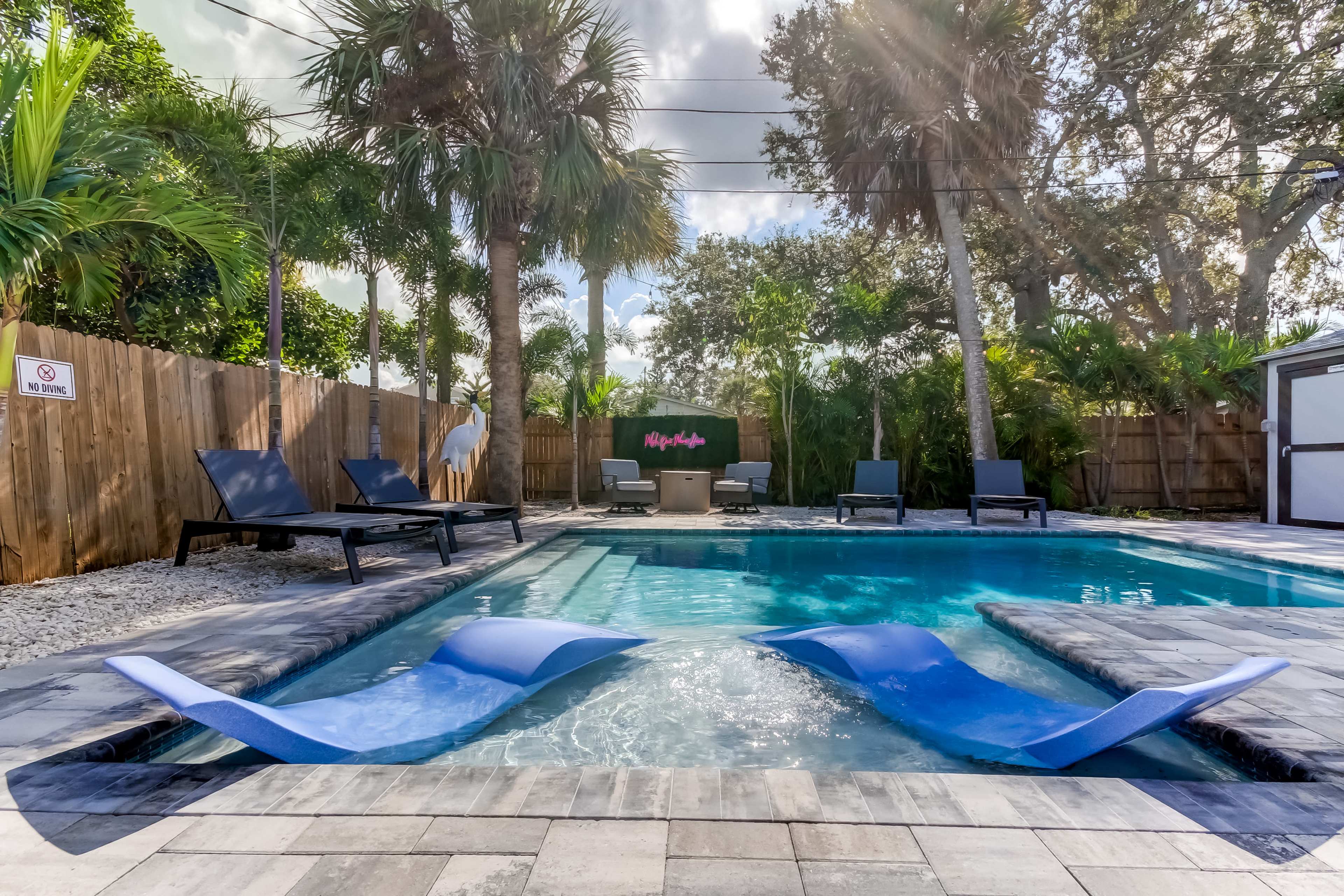 A blue swimming pool with lounge chairs and palm trees is surrounded by a stone patio and wooden fencing.