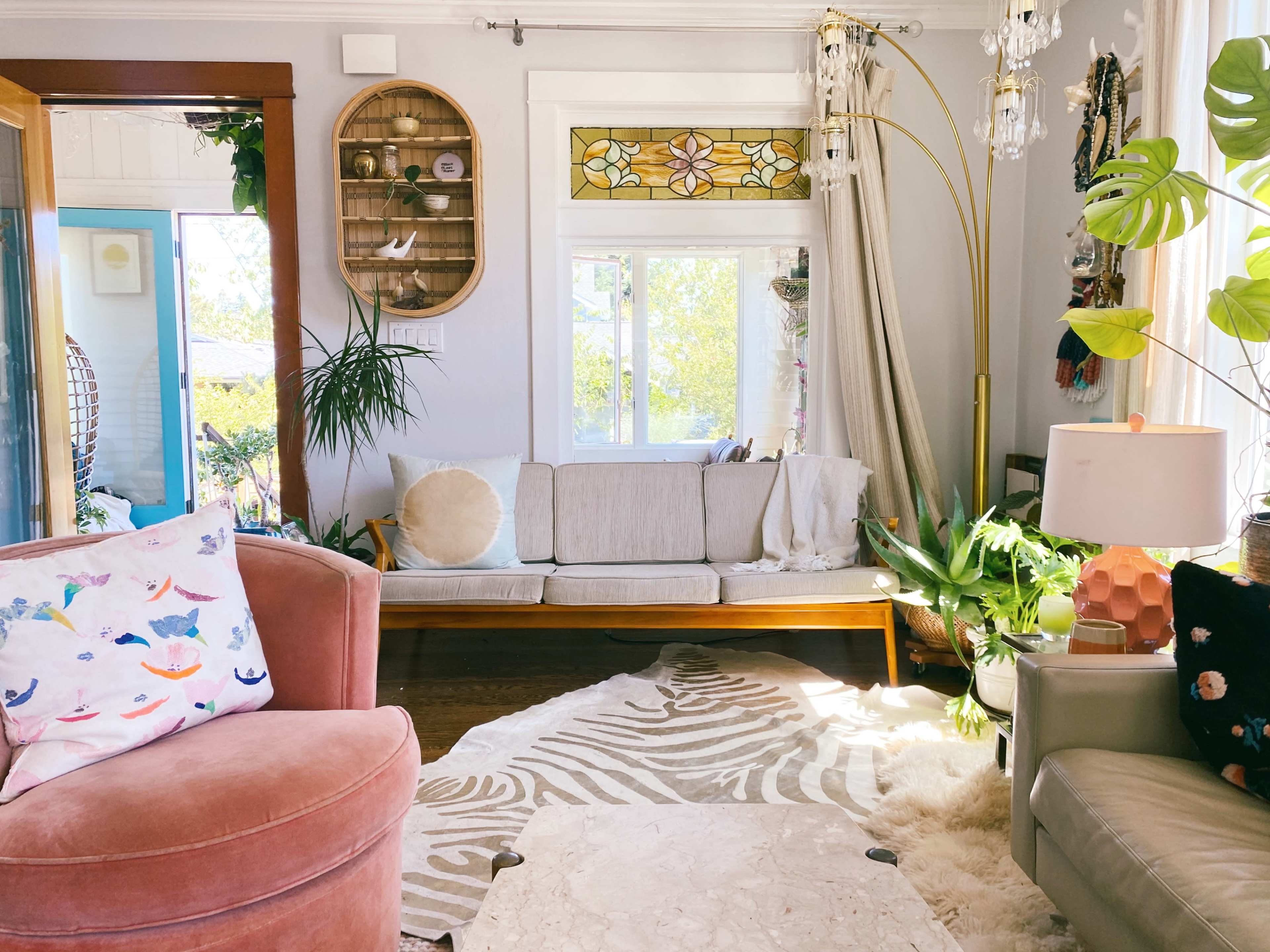 The living room features a light-colored couch, a pink armchair, and various plants, with a decorative rug and a stained glass window in the background.