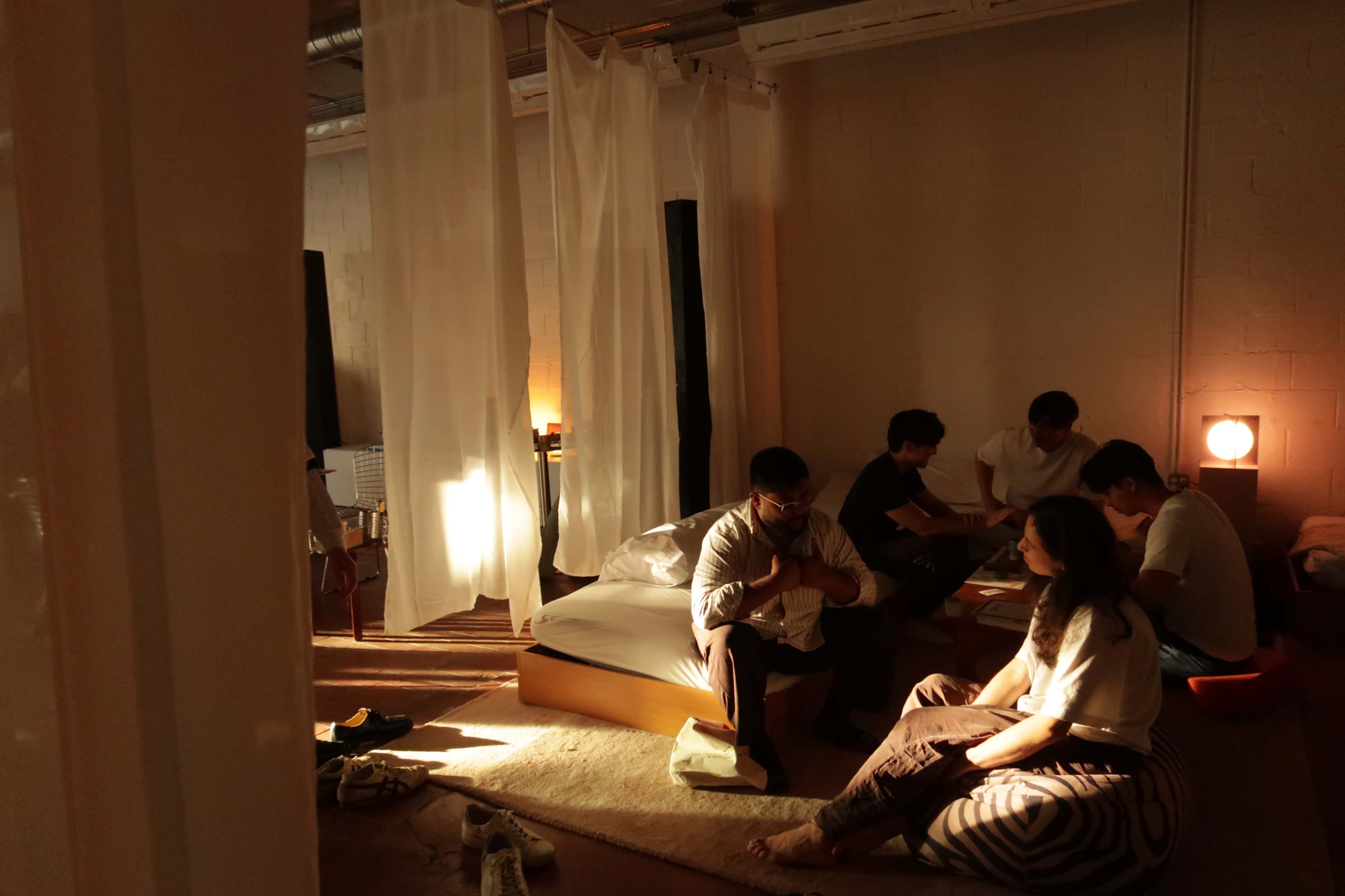 A group of five people sit and interact on the floor of a softly lit room, surrounded by white fabric drapes and various furniture.
