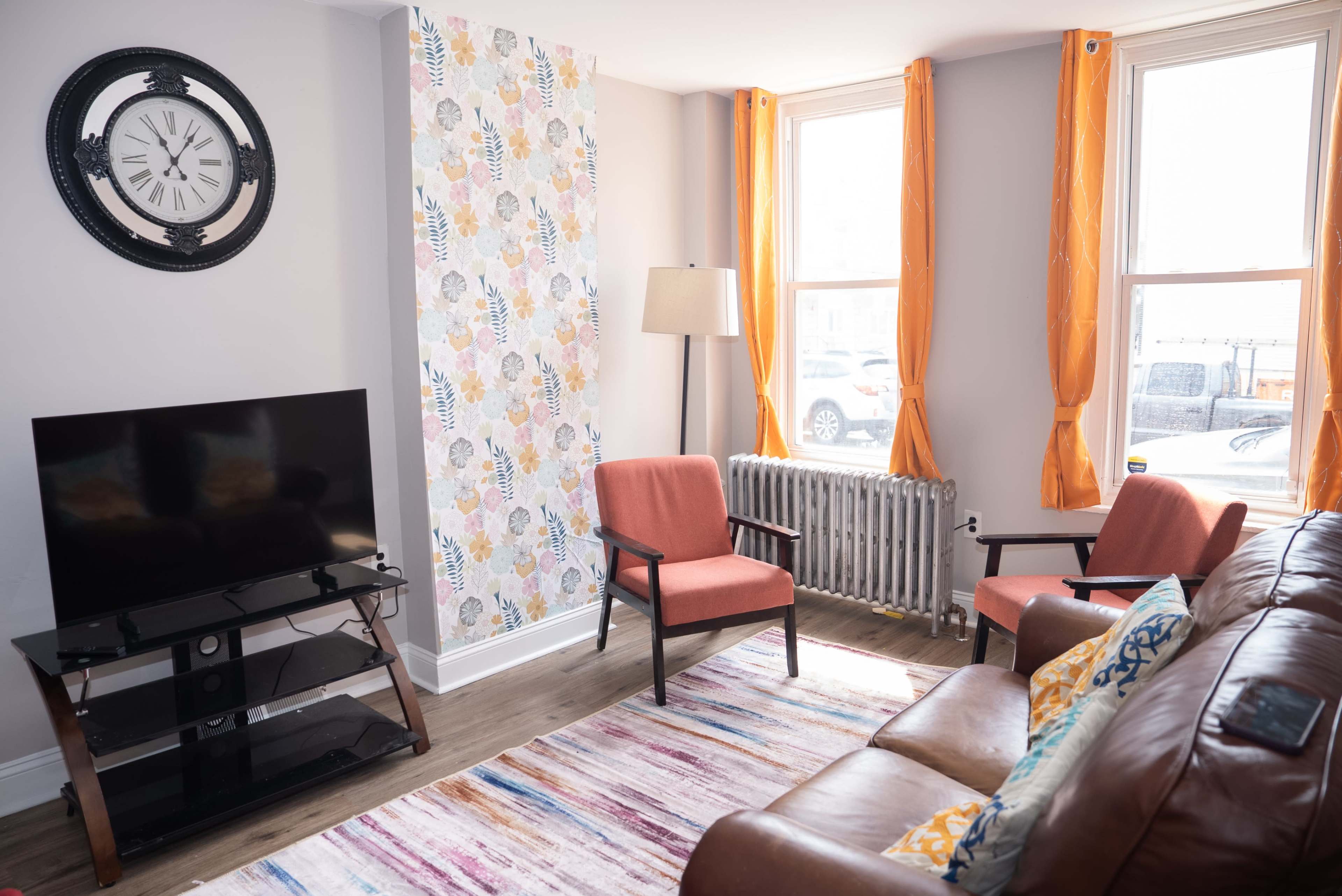 A living room features a television, two armchairs, a brown sofa, a patterned accent wall, and large windows with orange curtains.