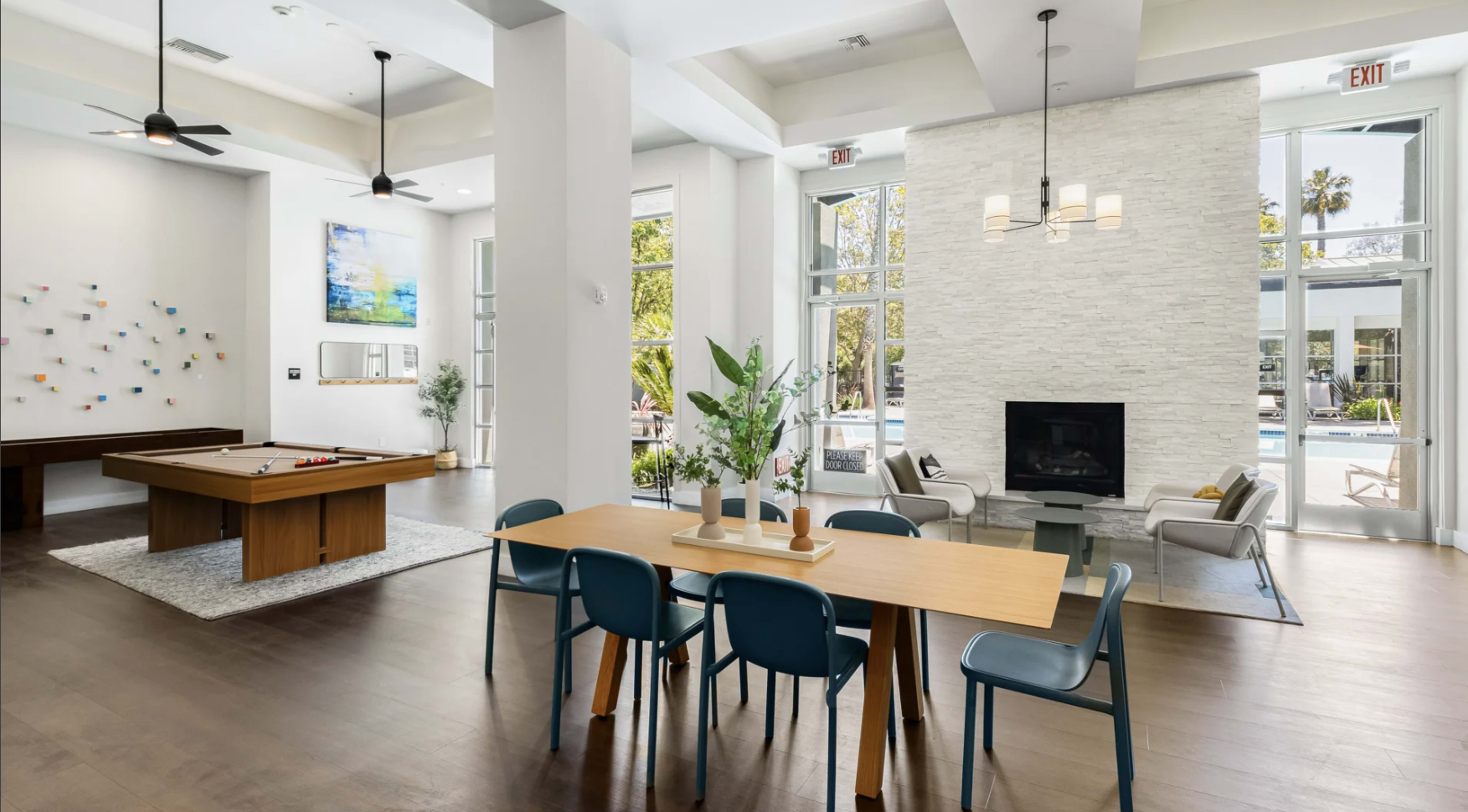 The image shows a modern indoor lounge area featuring a pool table, a dining table with blue chairs, and a cozy seating arrangement near a fireplace, all surrounded by large windows.