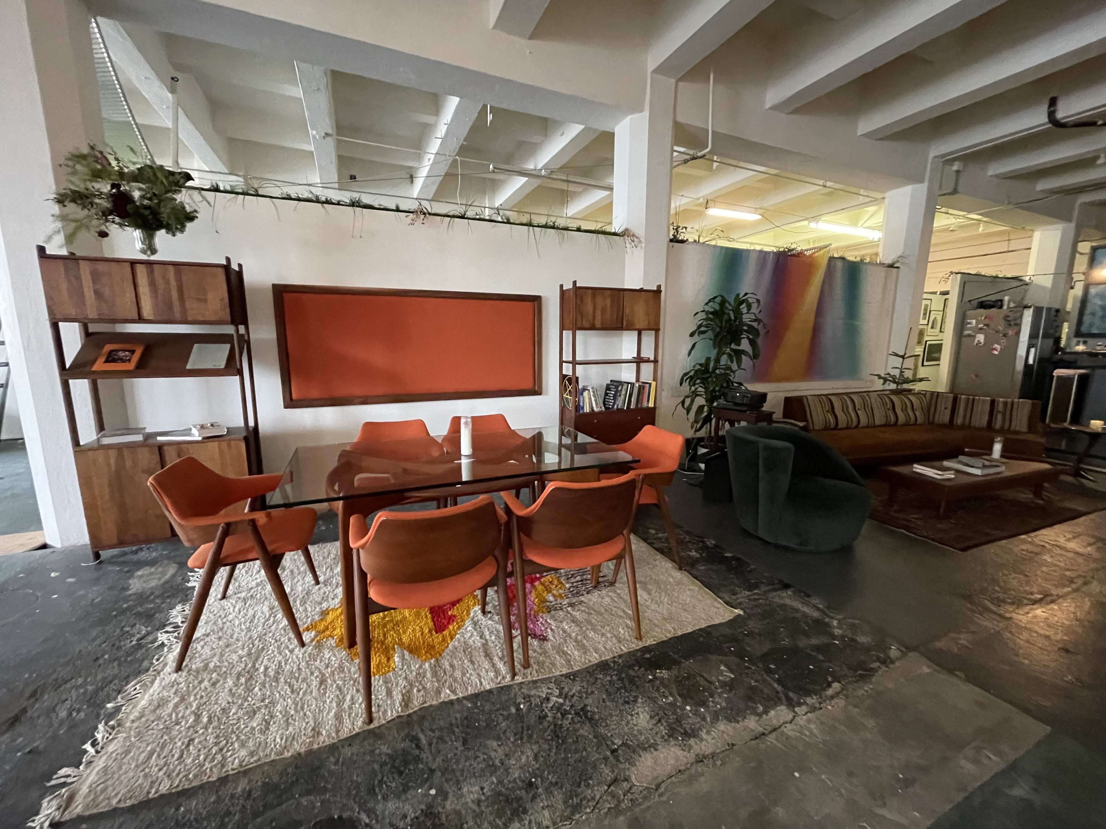 A modern open-plan office space featuring a glass dining table surrounded by orange chairs on a textured rug, a wooden bookshelf, and a cozy seating area with a green sofa and colorful artwork in the background.