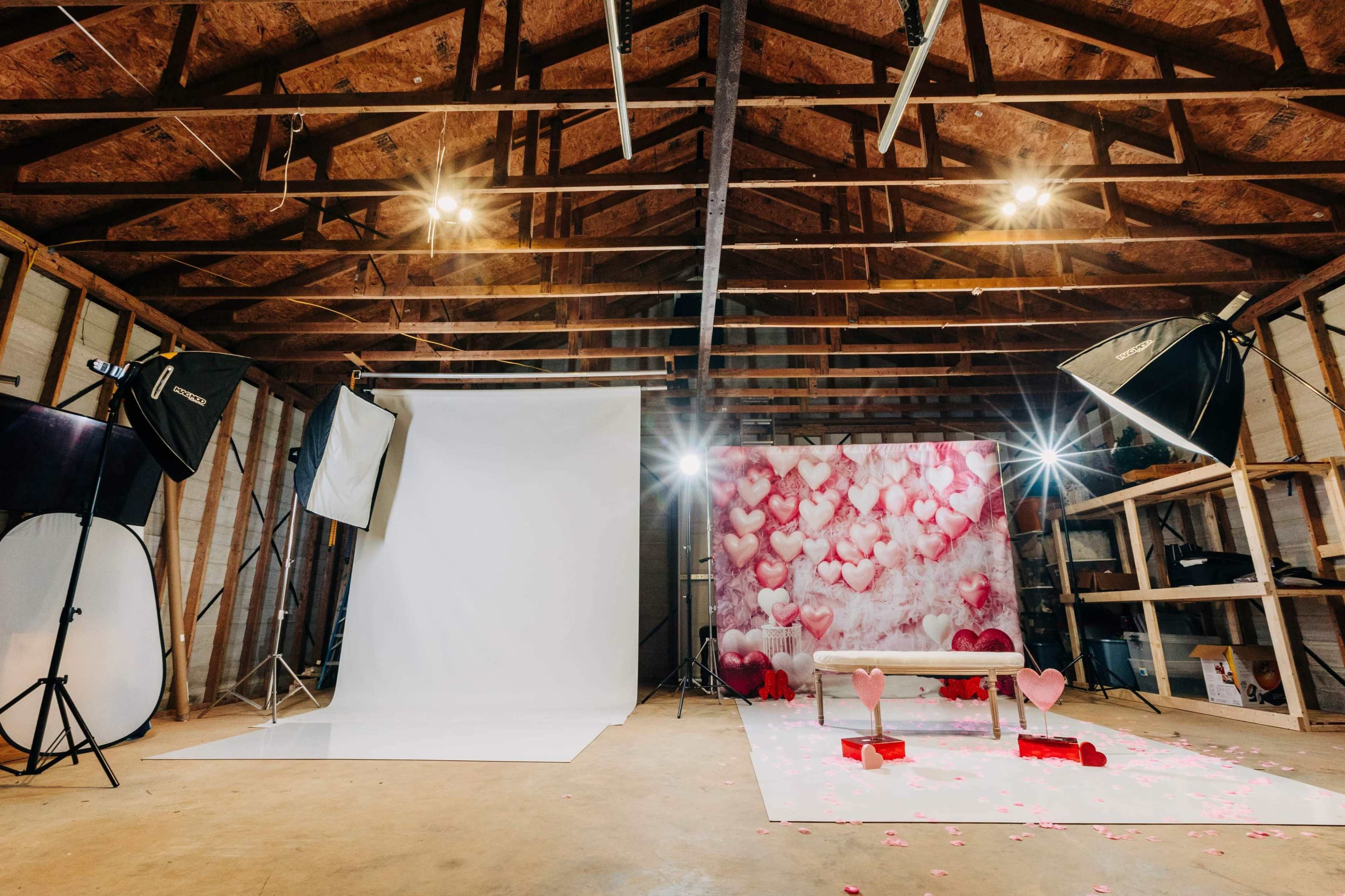 A photography studio featuring a white backdrop, a pink heart-themed wall, and lighting equipment arranged around a bench with decorative hearts.