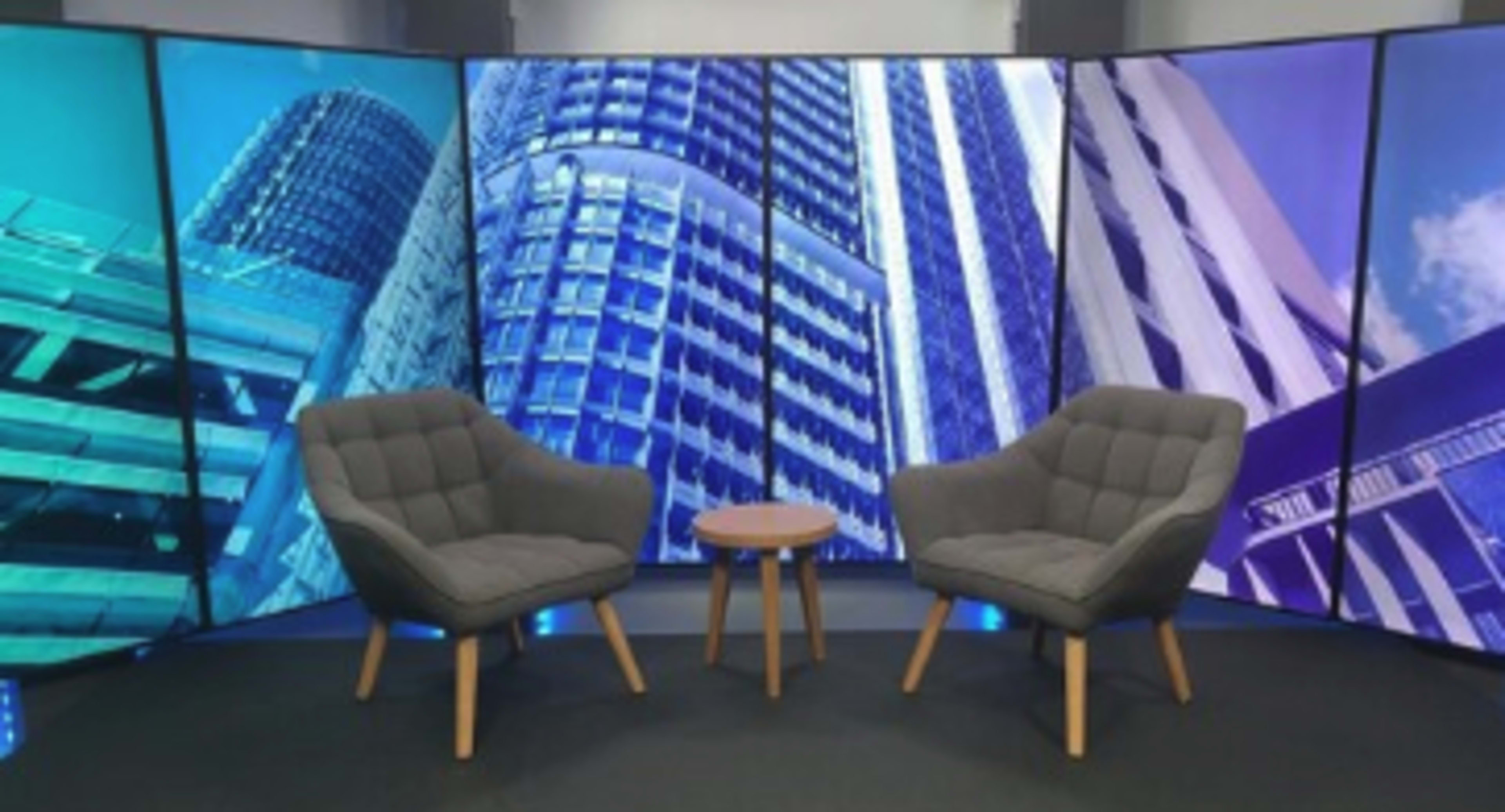 A modern studio set with two gray upholstered chairs facing a small wooden table, surrounded by backdrop panels featuring skyscraper images.