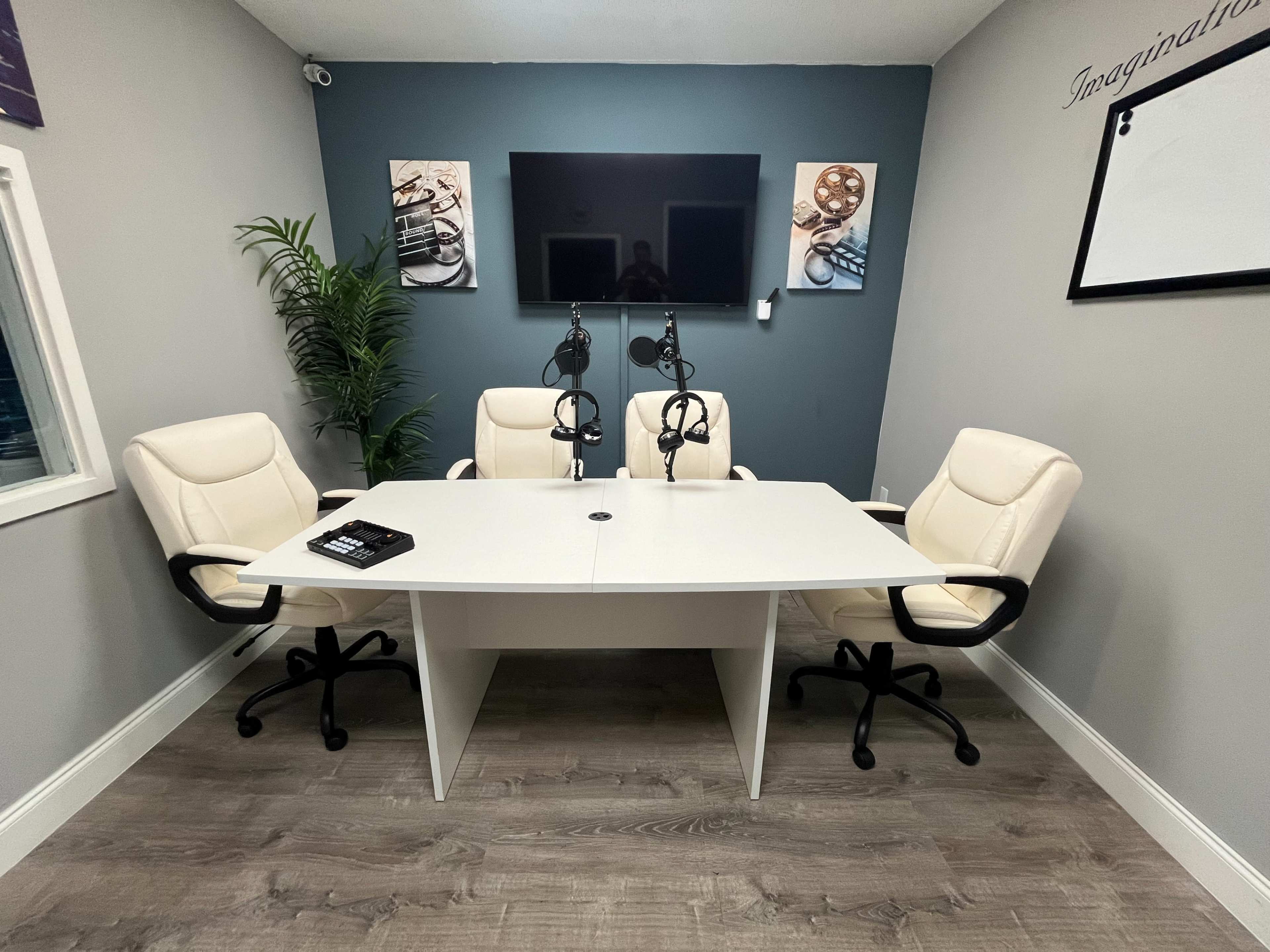 A modern conference room features a white table with four chairs, two of which have microphones attached, and a large television mounted on the wall.