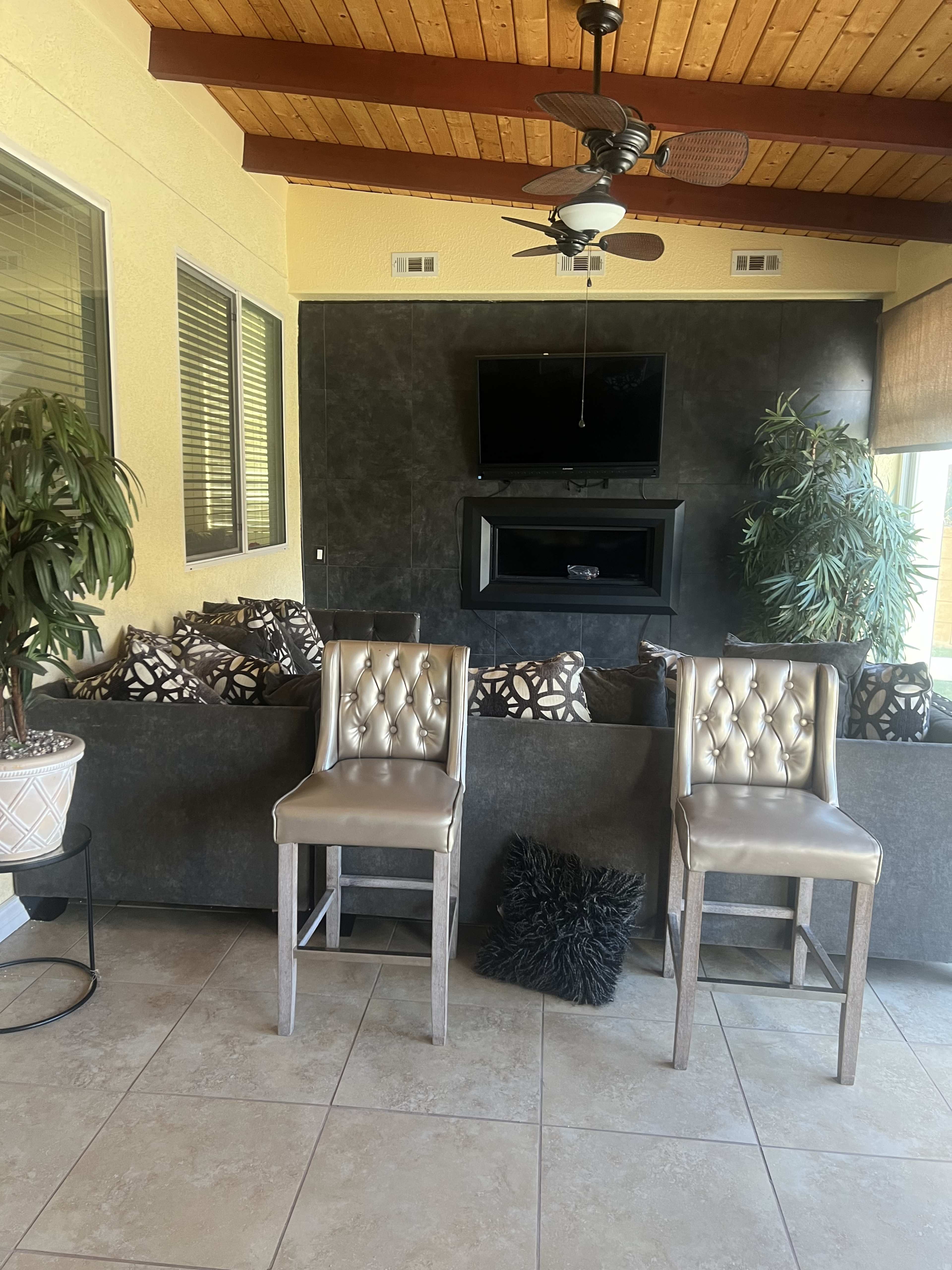 A cozy living space with two silver barstools facing a dark feature wall that has a mounted television and a modern fireplace, surrounded by plush seating and green plants.