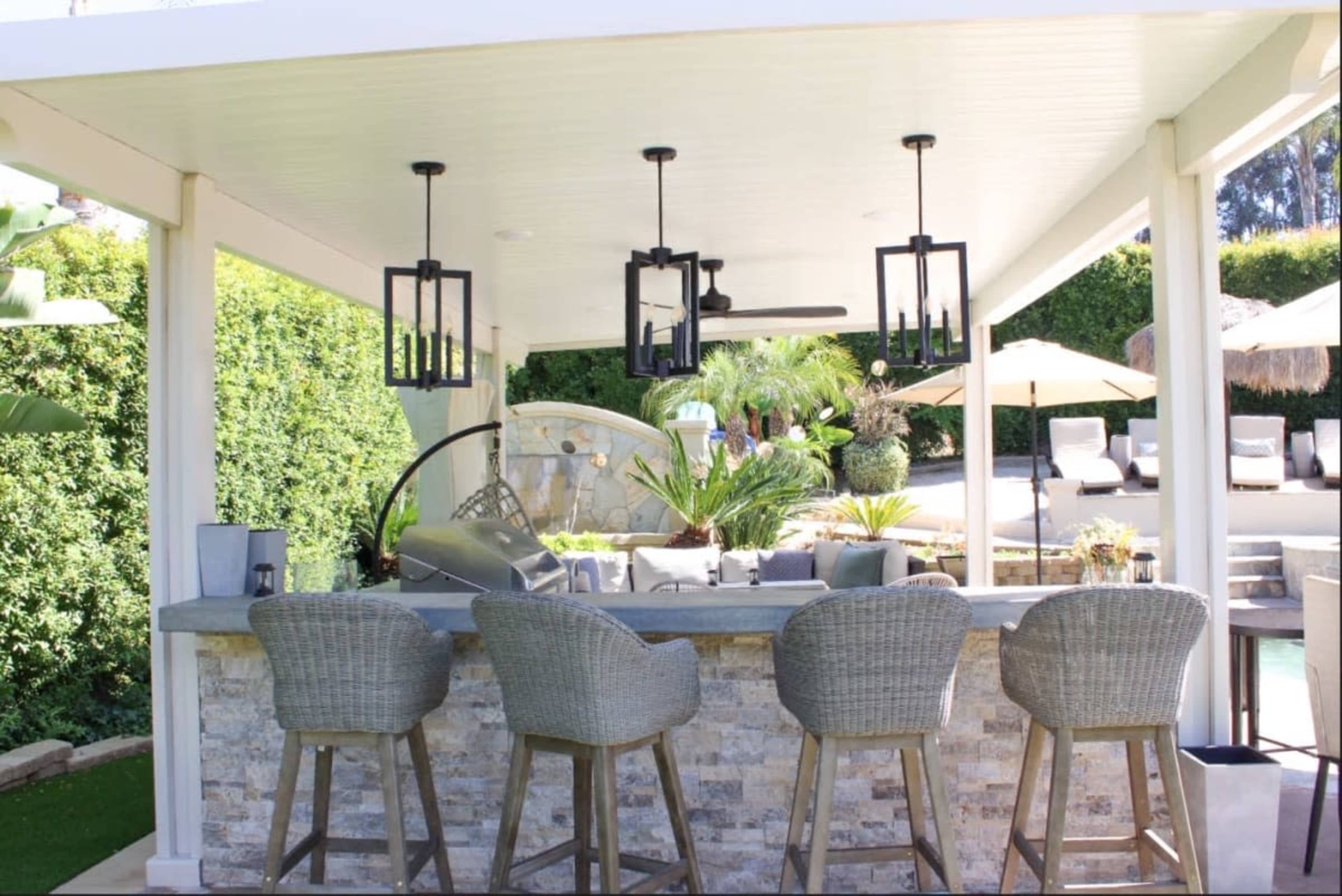 The image shows an outdoor bar area with stone countertops and four wicker bar stools, surrounded by greenery and lounge chairs.