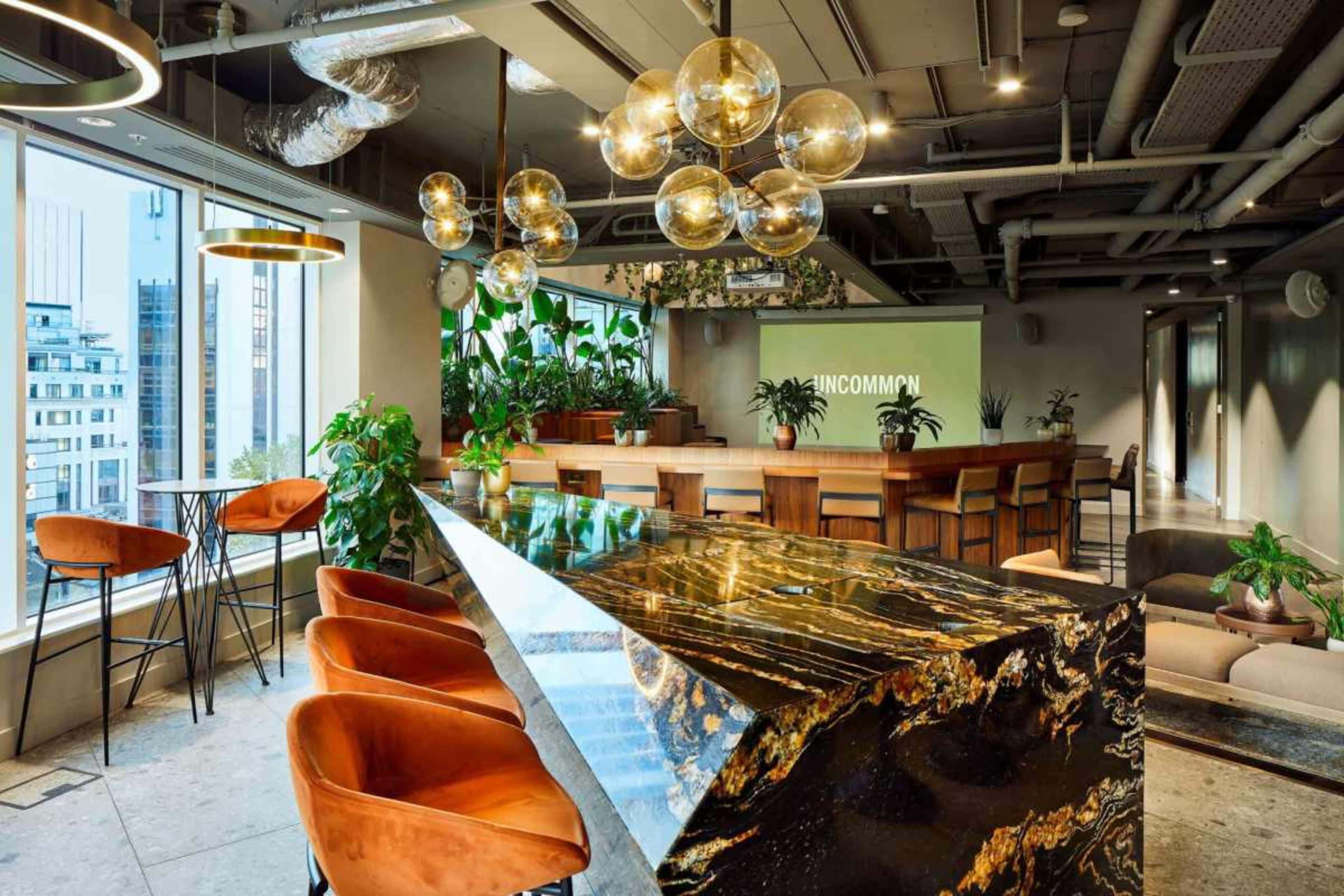 The image shows a modern office space with a sleek bar featuring a unique stone countertop, surrounded by plants and stylish seating arrangements.