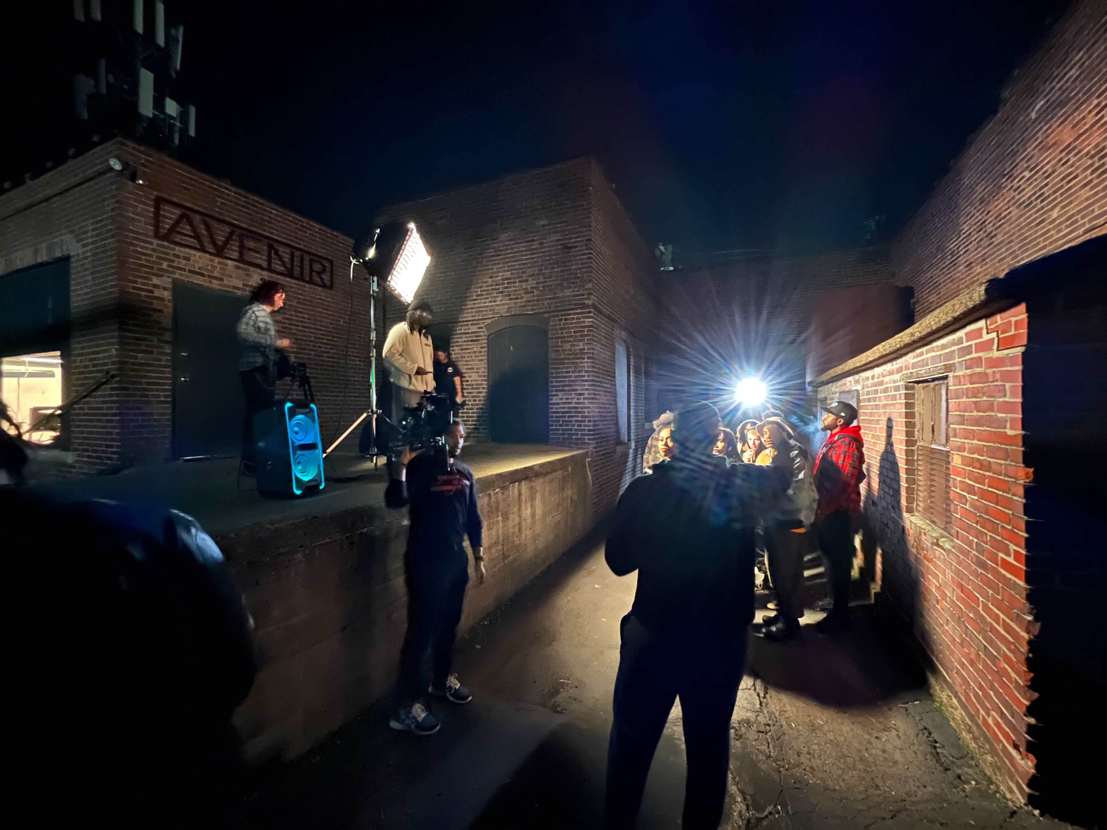 A group of people stands in a dimly lit outdoor area while a figure on a small platform speaks into a microphone, illuminated by bright lights.