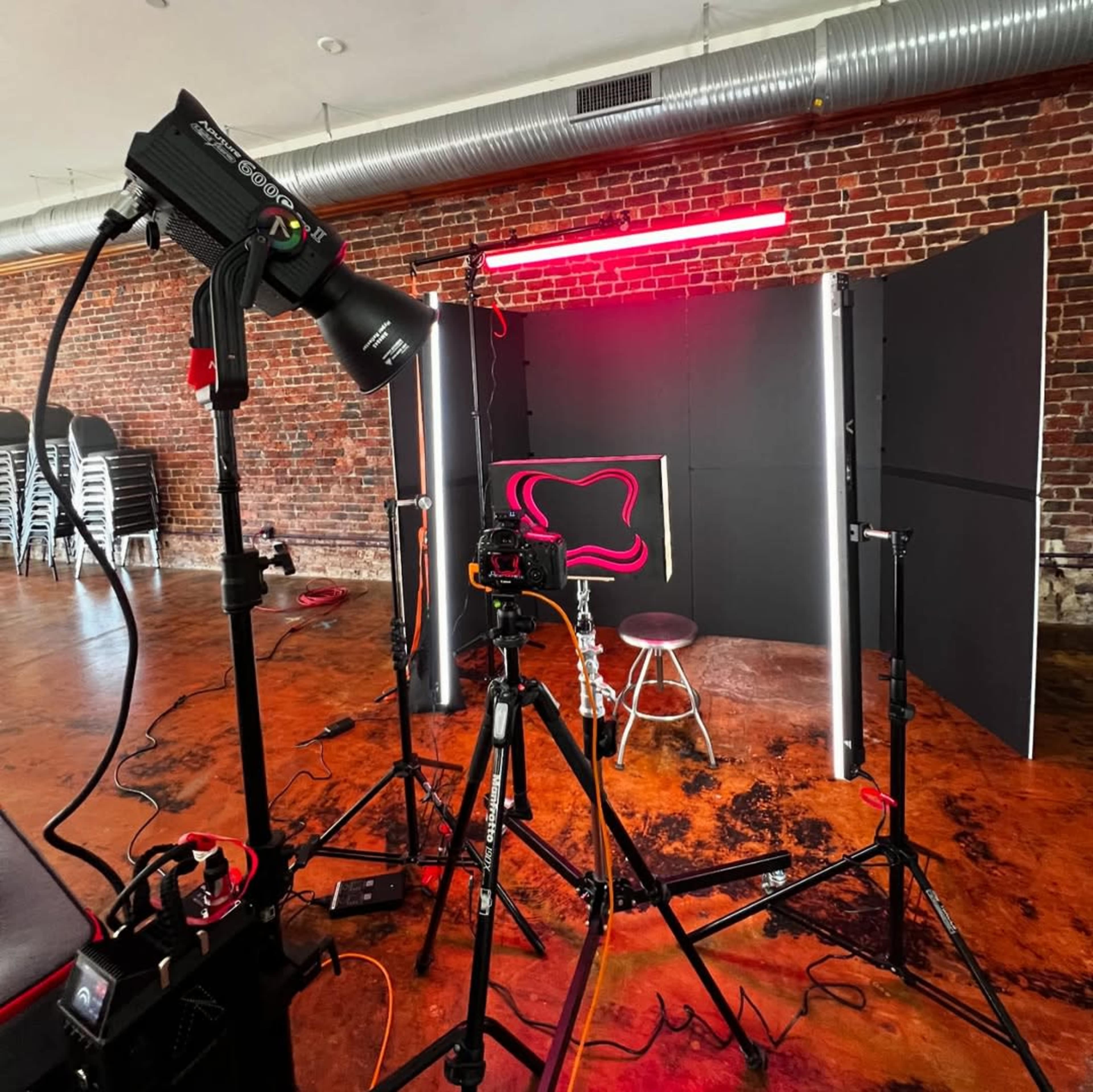 The image shows a photography setup with a tripod, lights, and a backdrop in a room featuring exposed brick walls and polished concrete flooring.