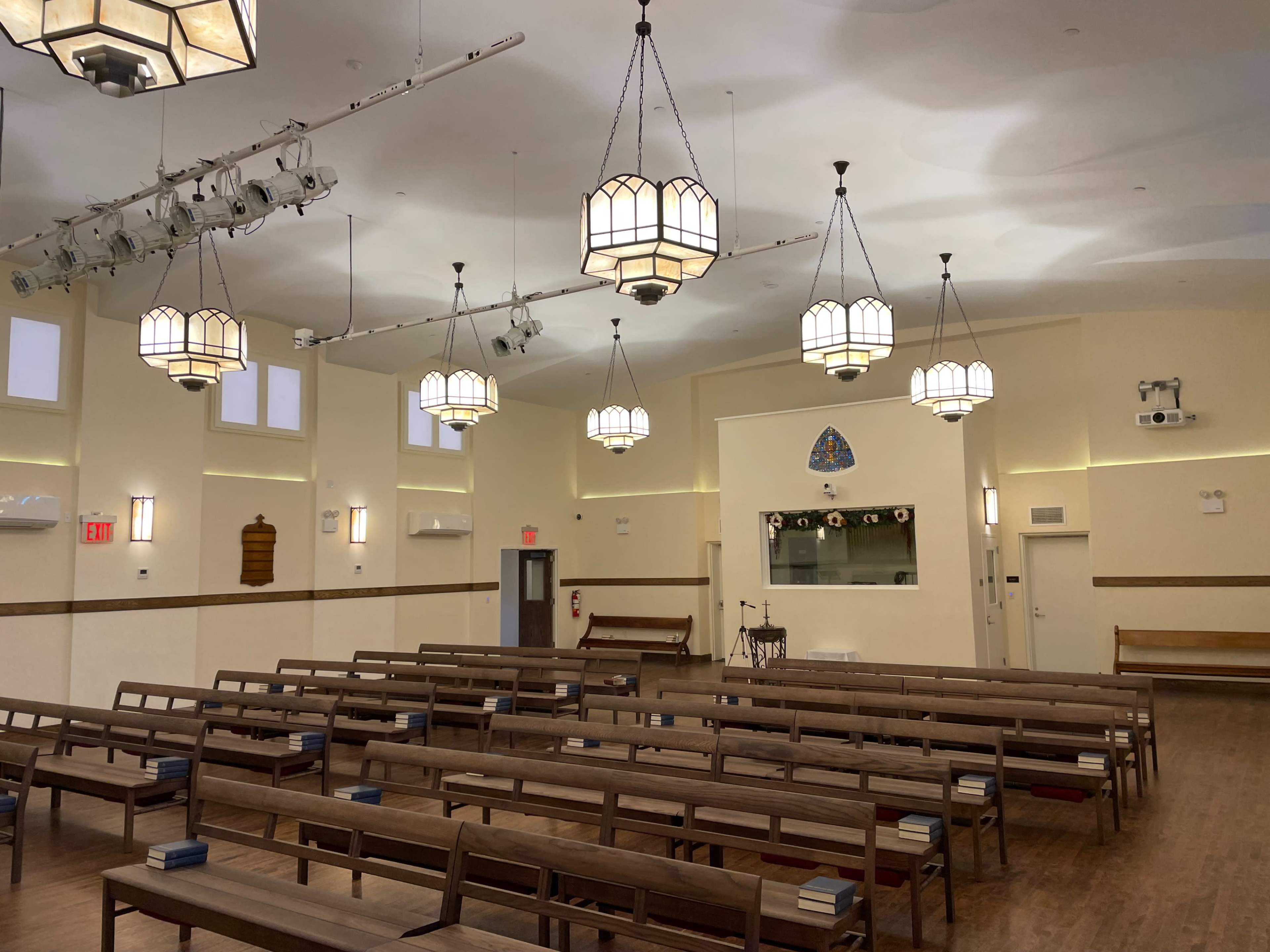 The interior of a hall with wooden benches arranged in rows, pendant lights hanging from the ceiling, and a small stage area at the front.