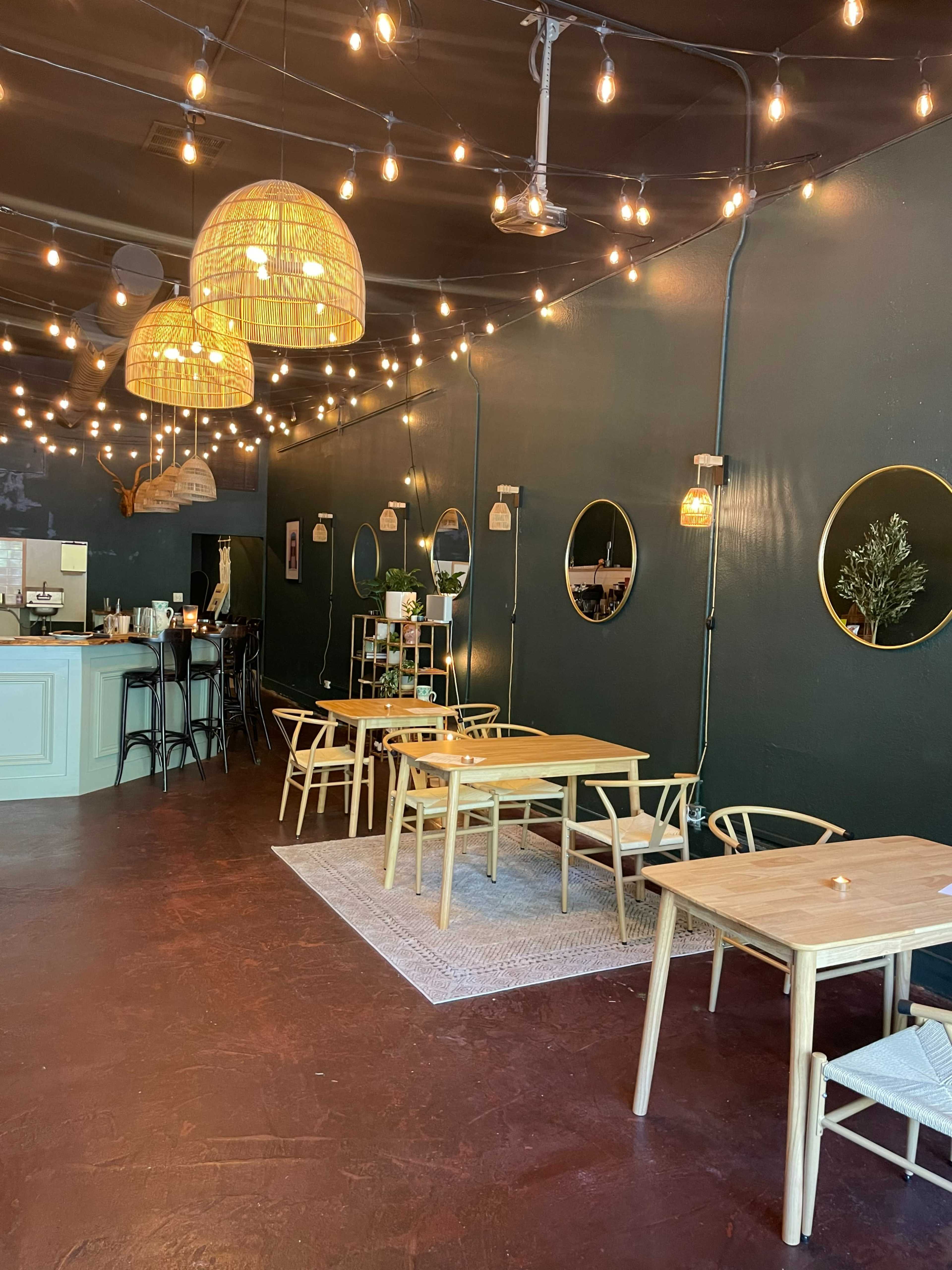 A cozy cafe interior with wooden tables, circular mirrors, and string lights hanging from a dark green wall.