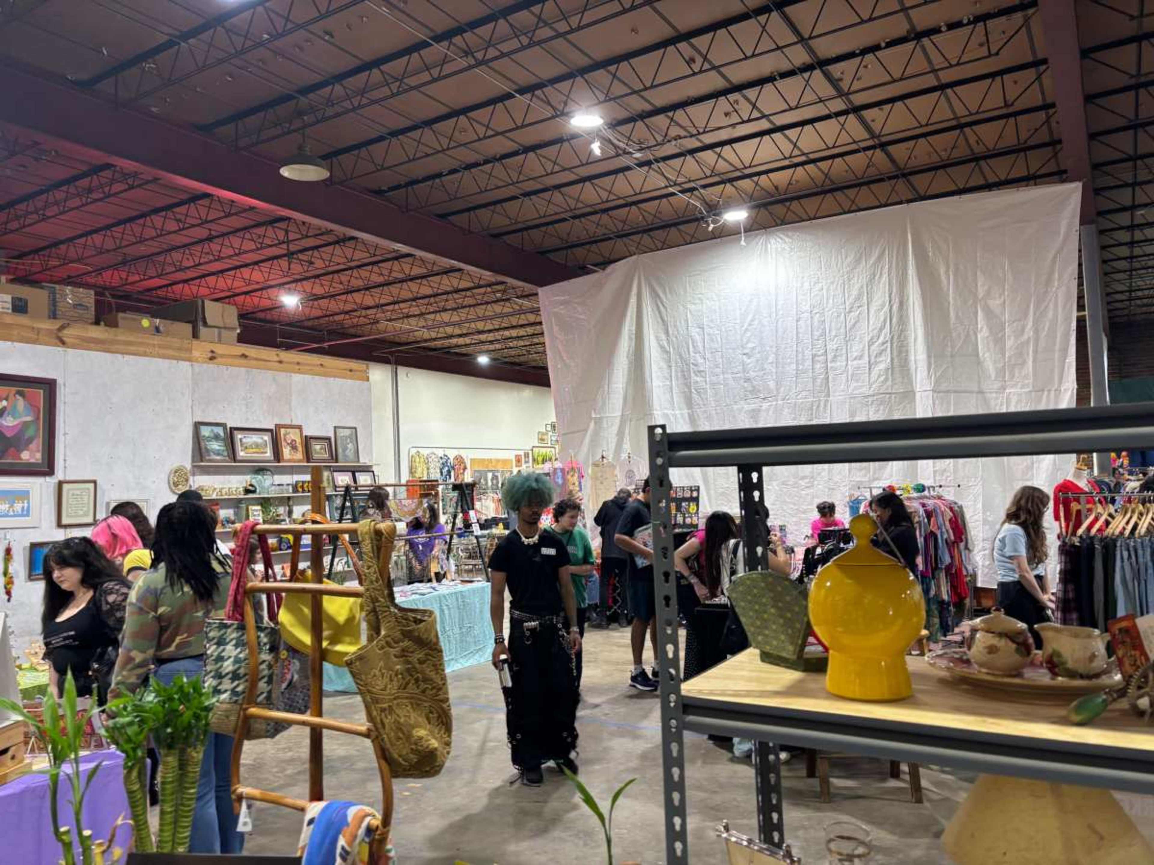 The image shows people browsing and shopping in a bustling indoor market or fair, with various stalls displaying clothing, crafts, and home decor items.