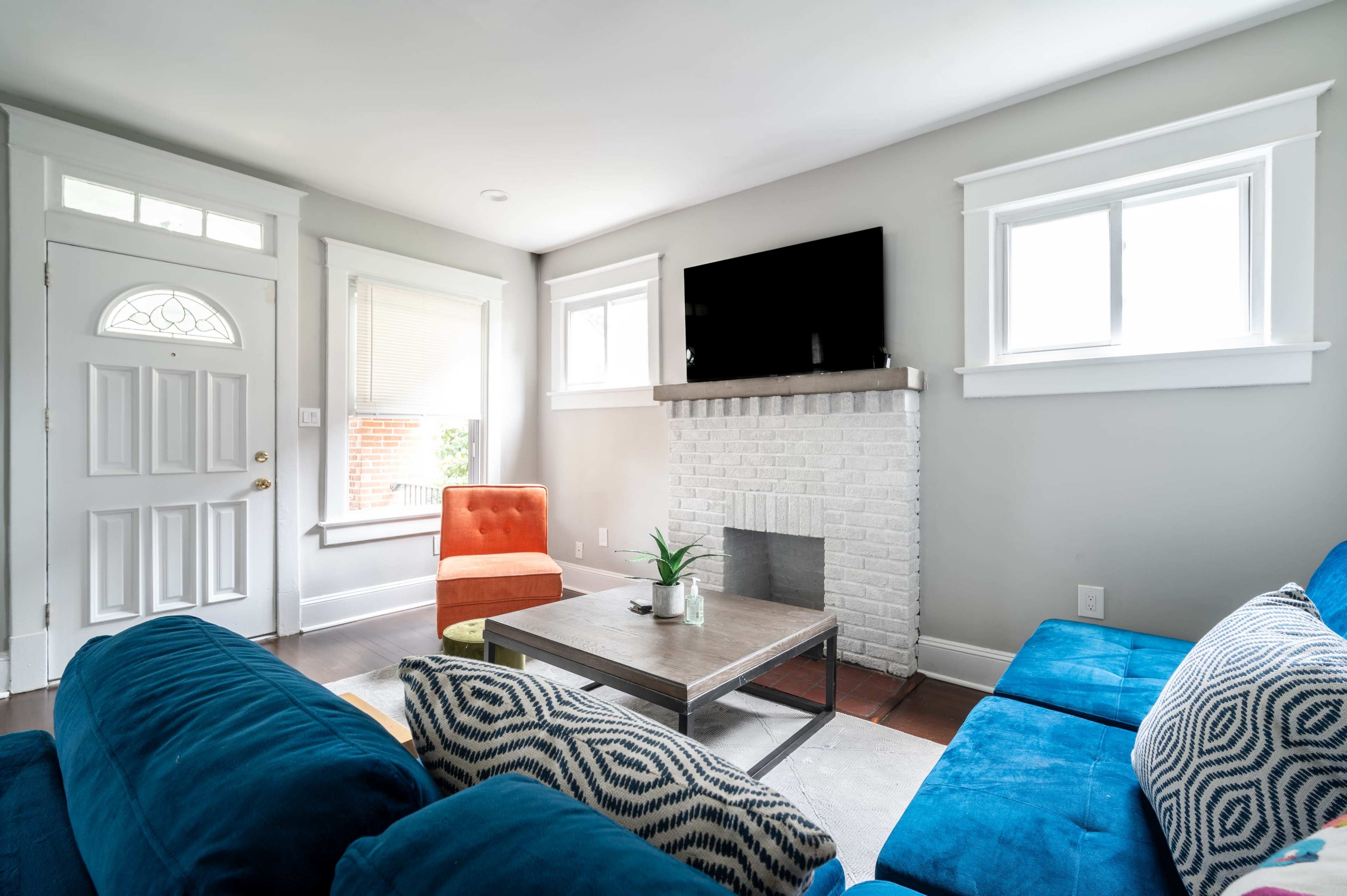 A living room with a blue sofa, an orange armchair, a square coffee table, and a white brick fireplace, all under a mounted television.