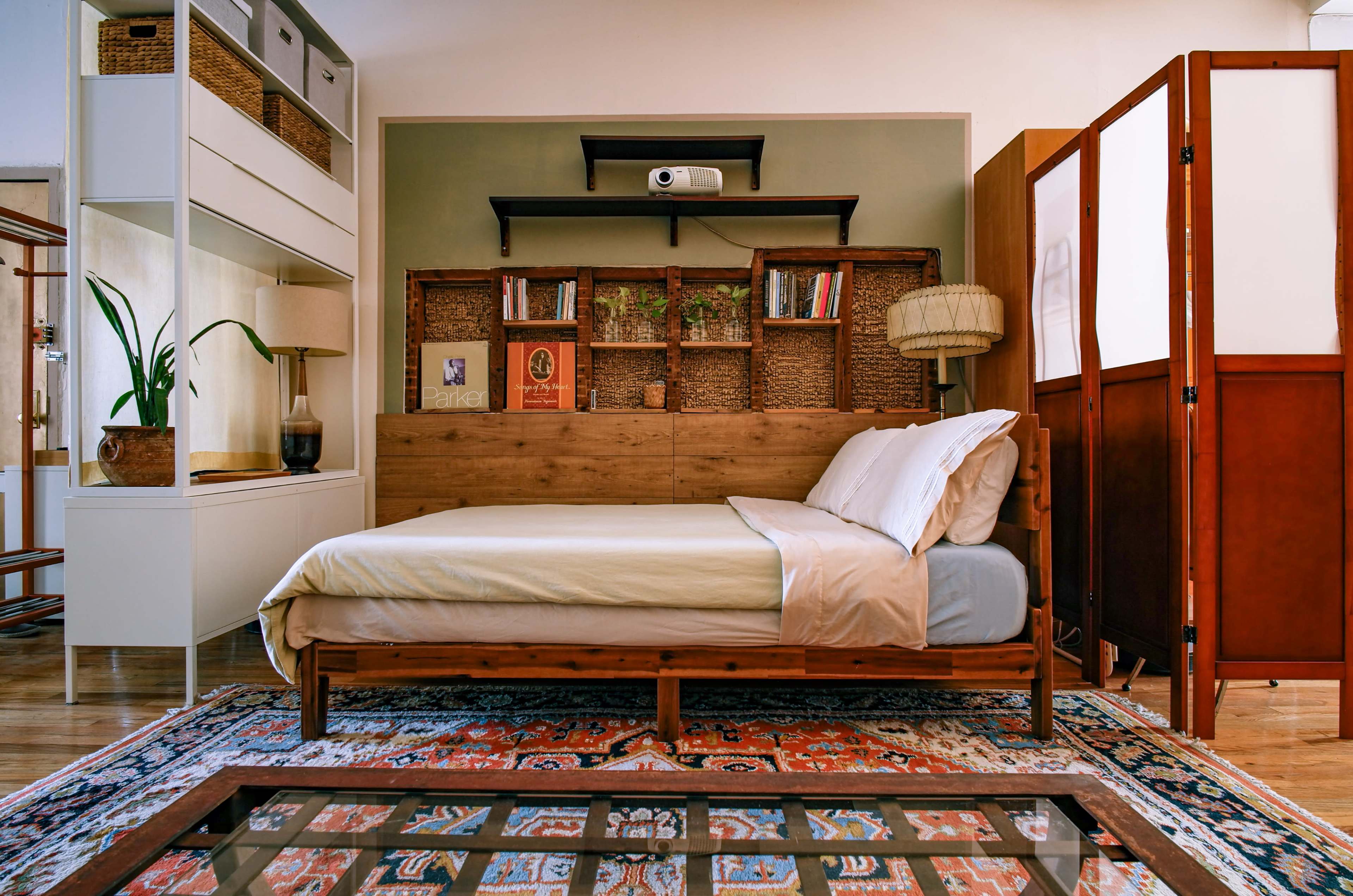 The image shows a neatly arranged bedroom featuring a wooden bed, a mounted projector, a bookshelf, and a decorative room divider.