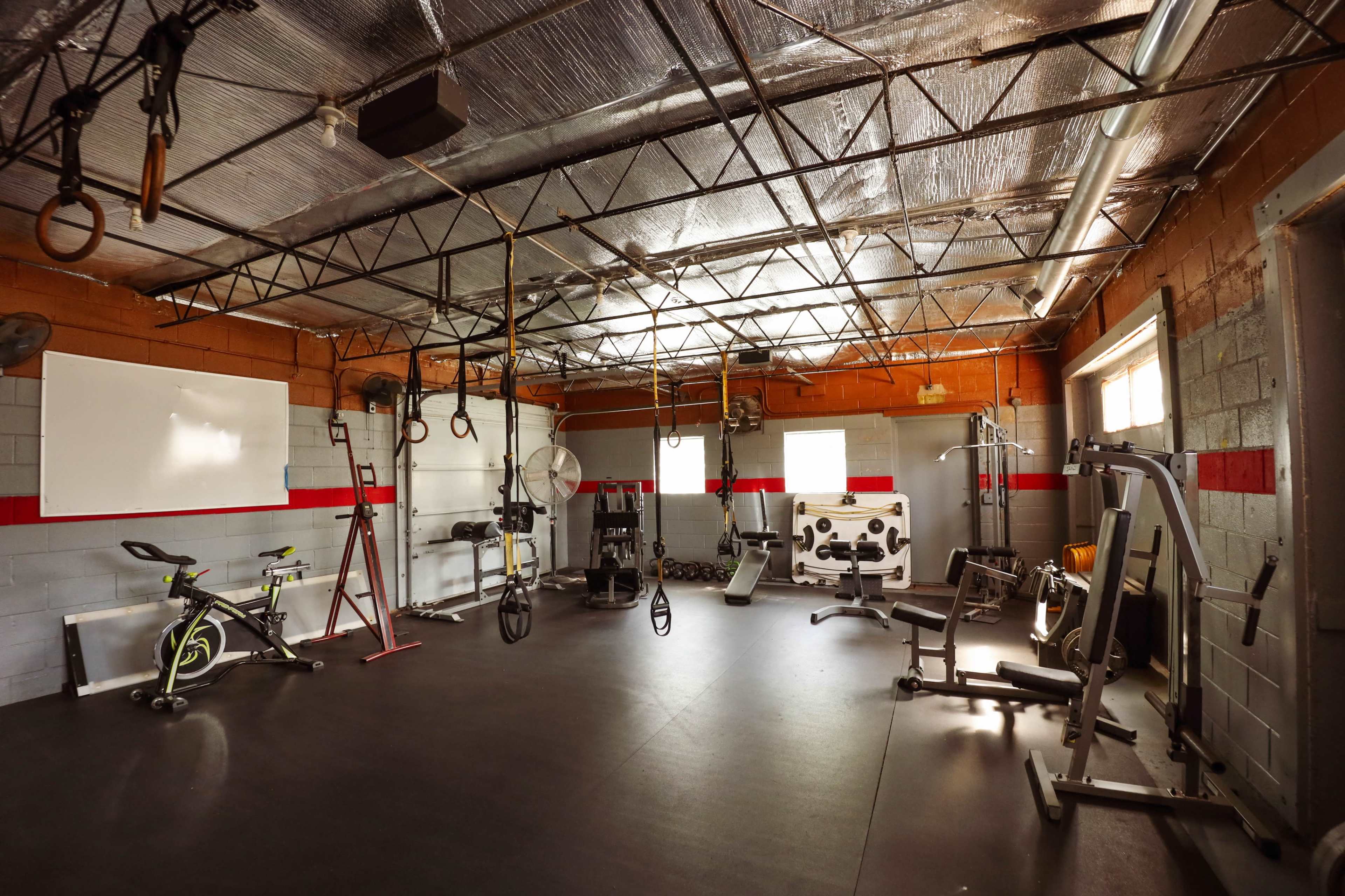 An empty gym with various exercise equipment, including a stationary bike, weight bench, and hanging straps, set against bare walls and exposed beams.
