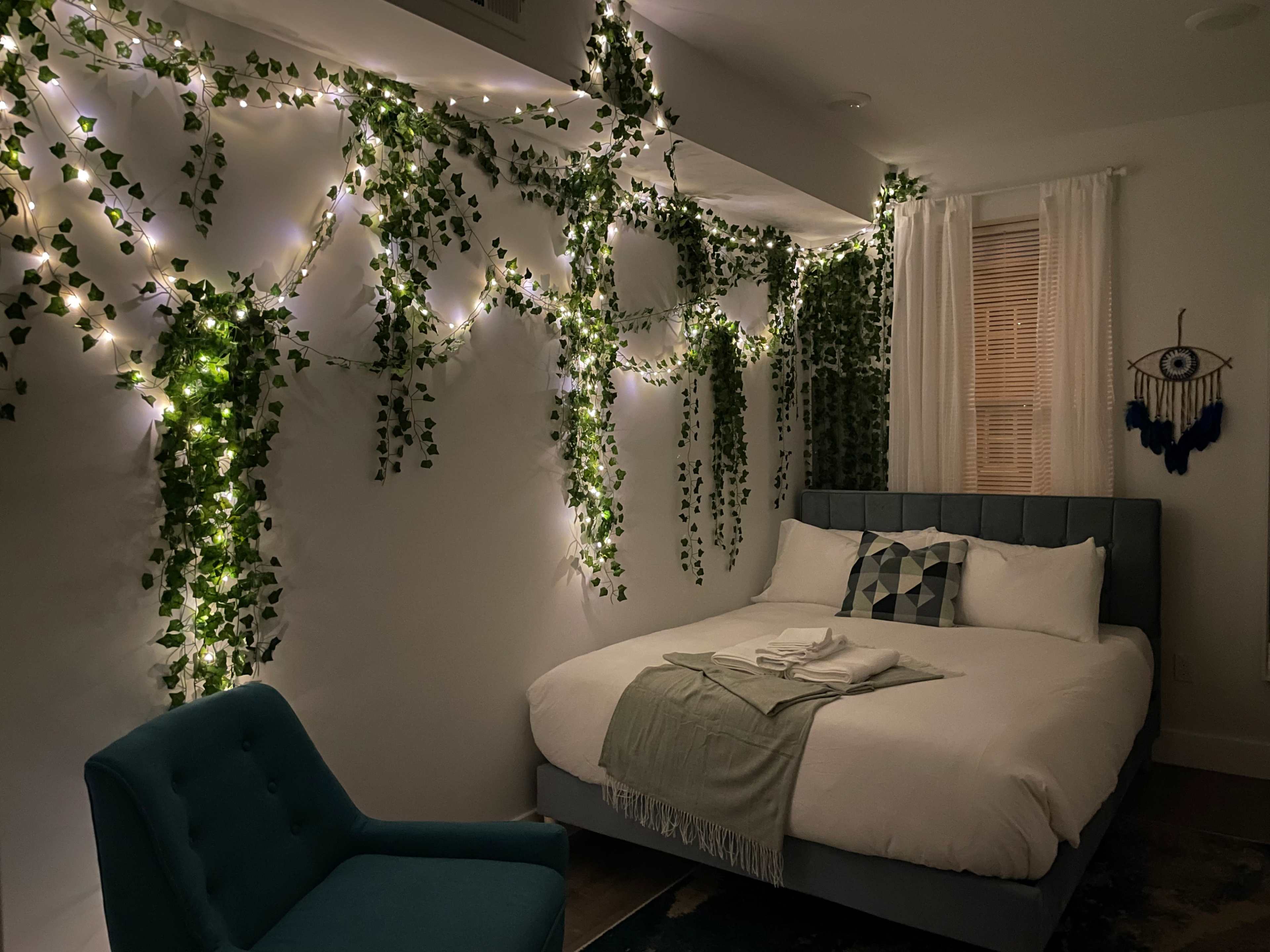 A bedroom with a bed, decorated wall with green vines and string lights, a blue chair, and a window covered with sheer curtains.