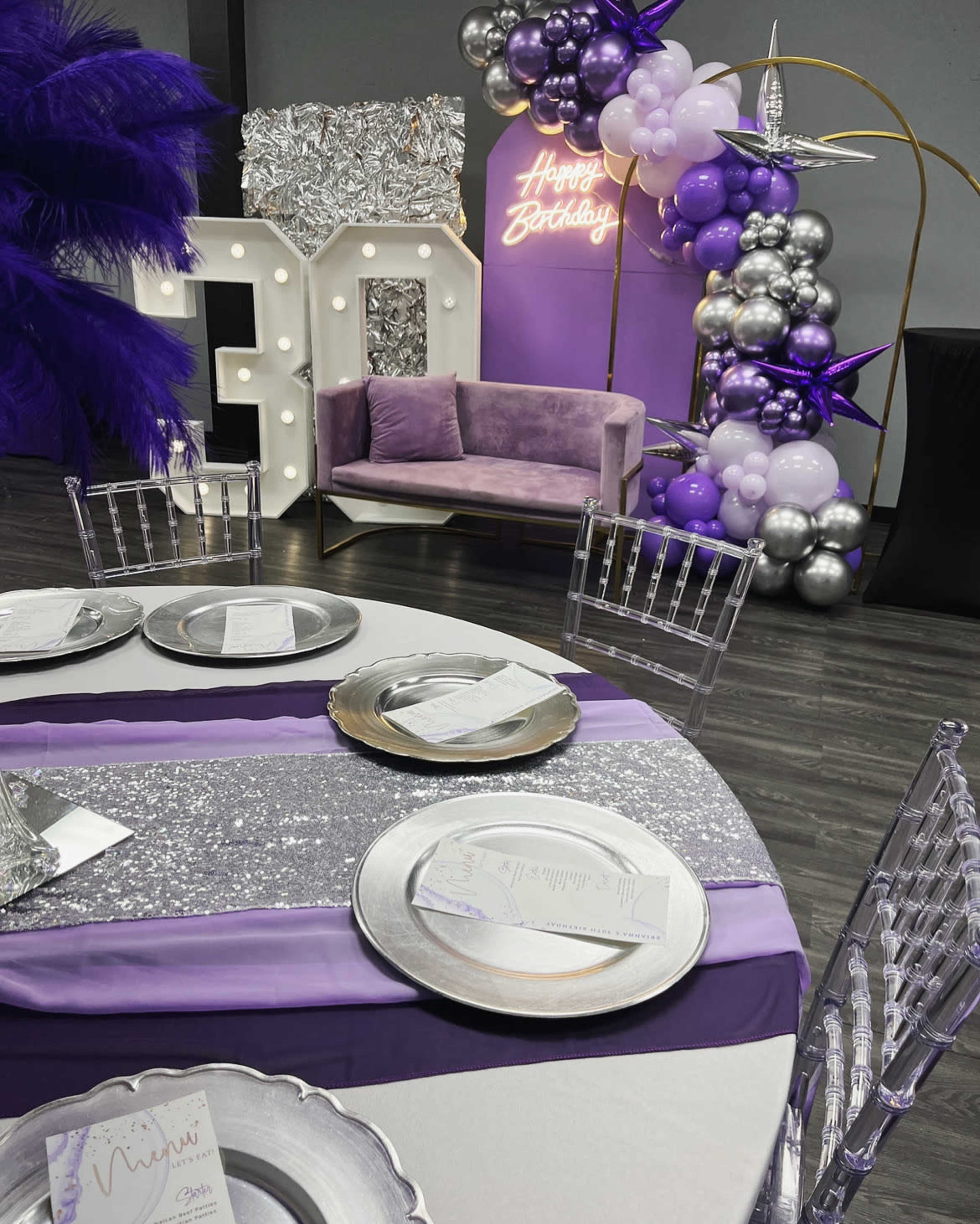 A decorated event space with a purple and silver color scheme, featuring a table set for a celebration, a plush sofa, and a balloon arch that says "Happy Birthday."