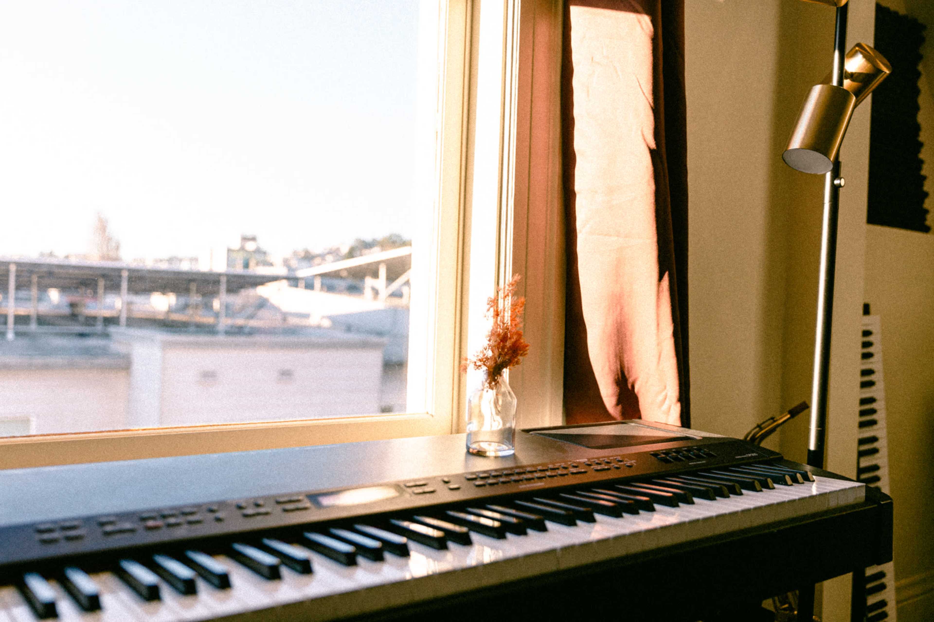 A keyboard sits next to a window, with a small vase of dried flowers in front and a lamp illuminating the scene.