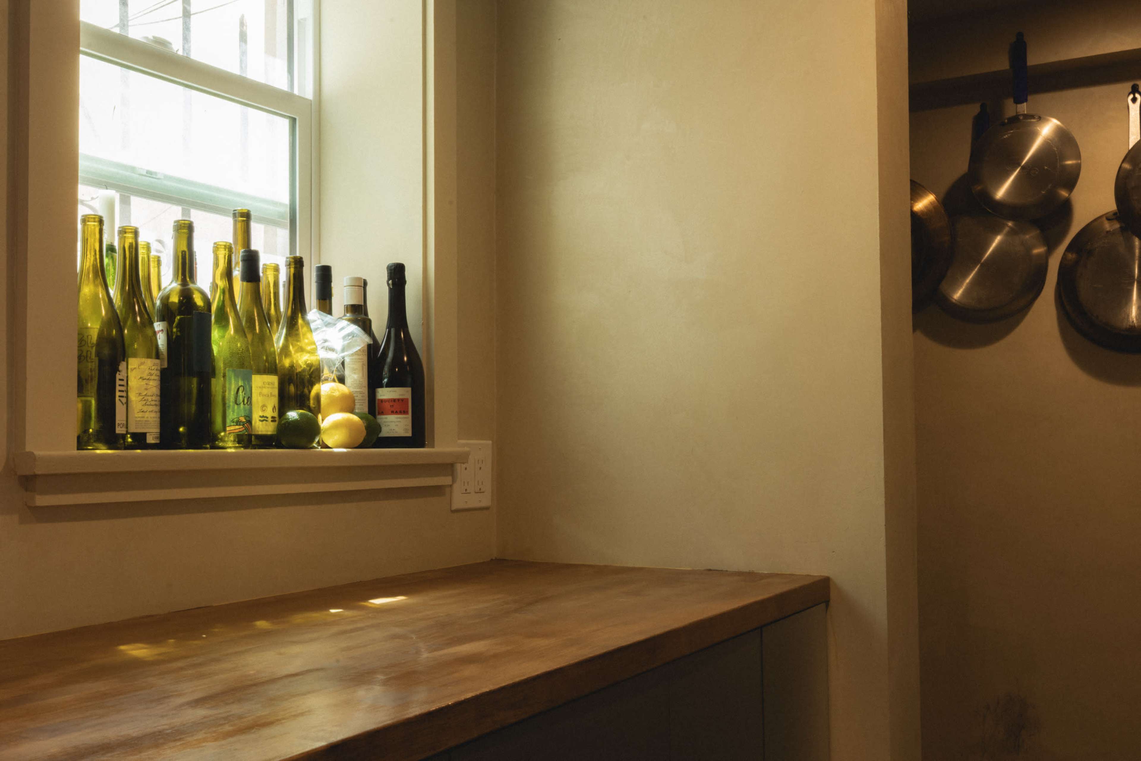 A row of empty wine bottles is arranged on the windowsill beside a bowl of lemons in a kitchen nook with a wooden countertop.