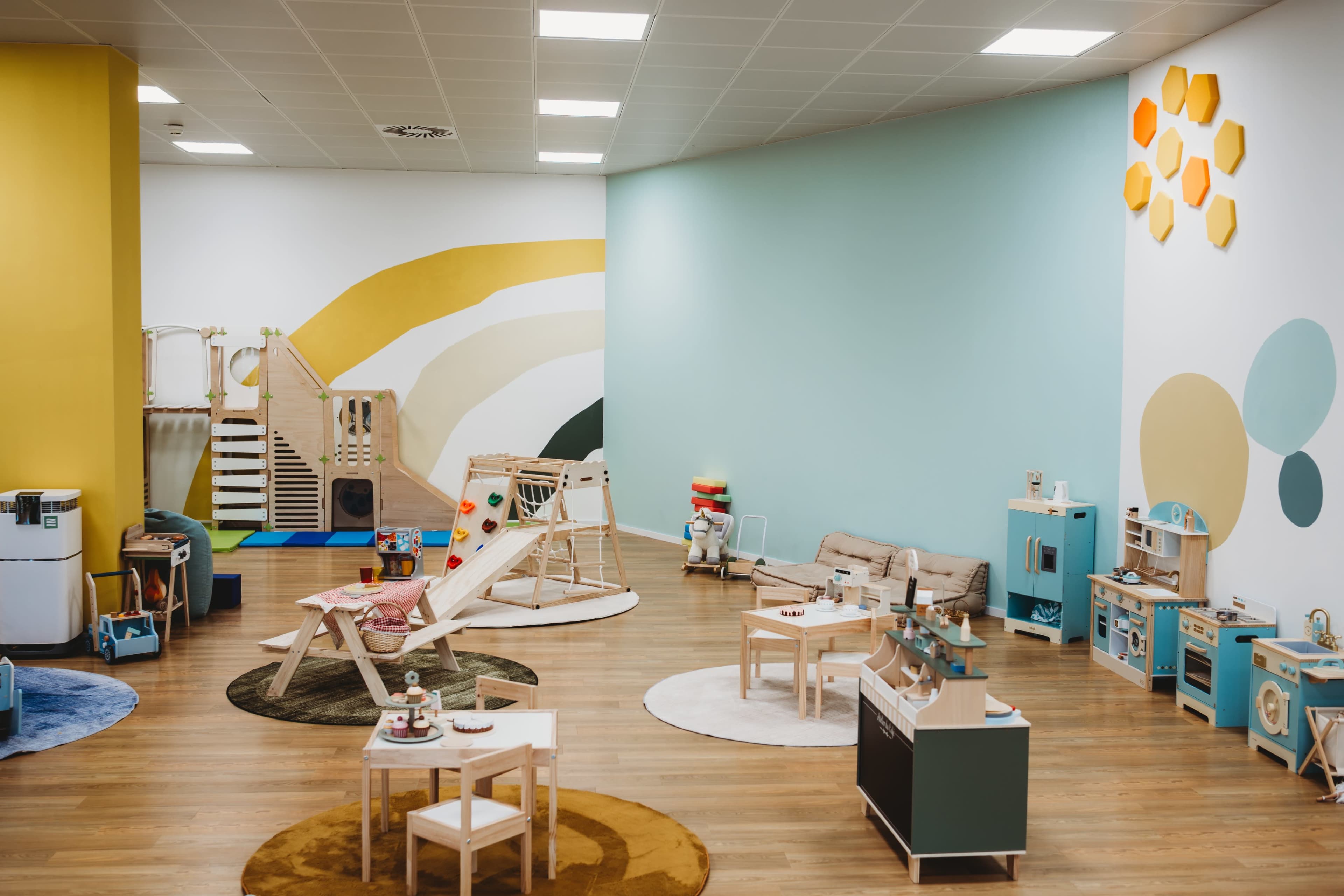 The image shows a brightly colored playroom equipped with various toys, furniture, and climbing structures, arranged on wooden flooring.