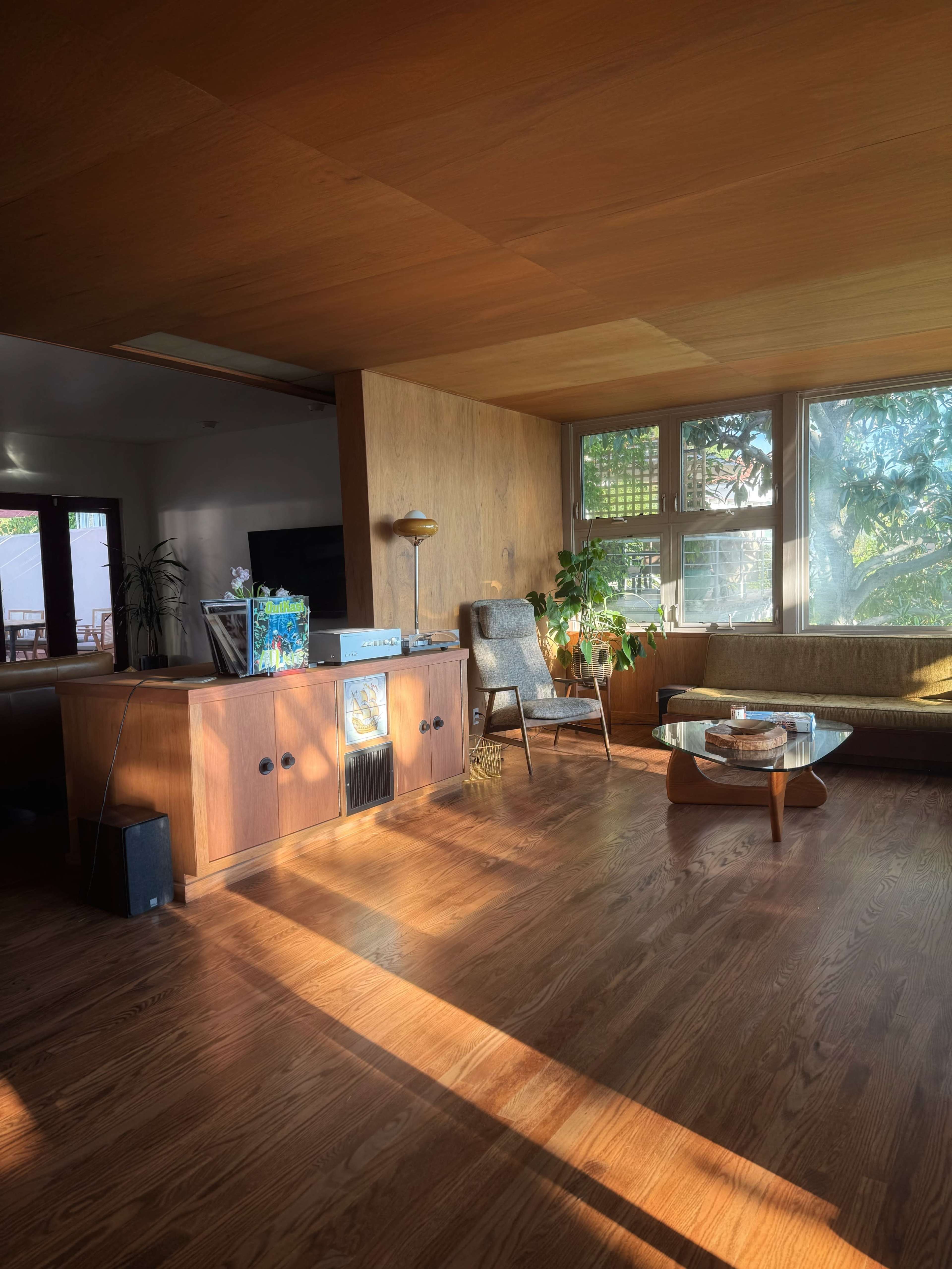 The image shows a sunlit living room with wooden walls, a mid-century modern armchair, and a low coffee table, alongside a large window that offers a view of greenery outside.