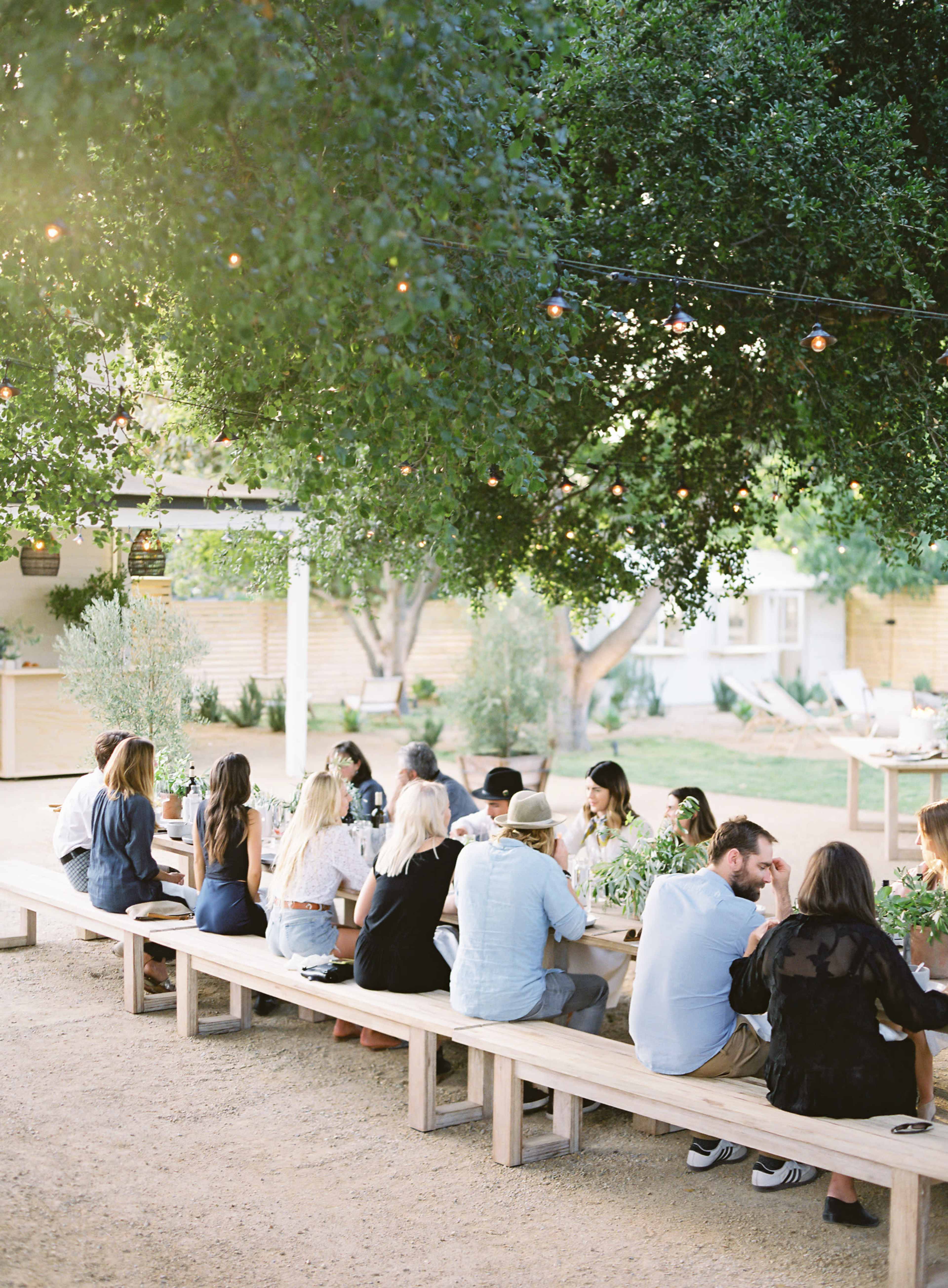 A group of people is seated at long wooden benches under trees with string lights, engaged in a gathering at an outdoor venue.
