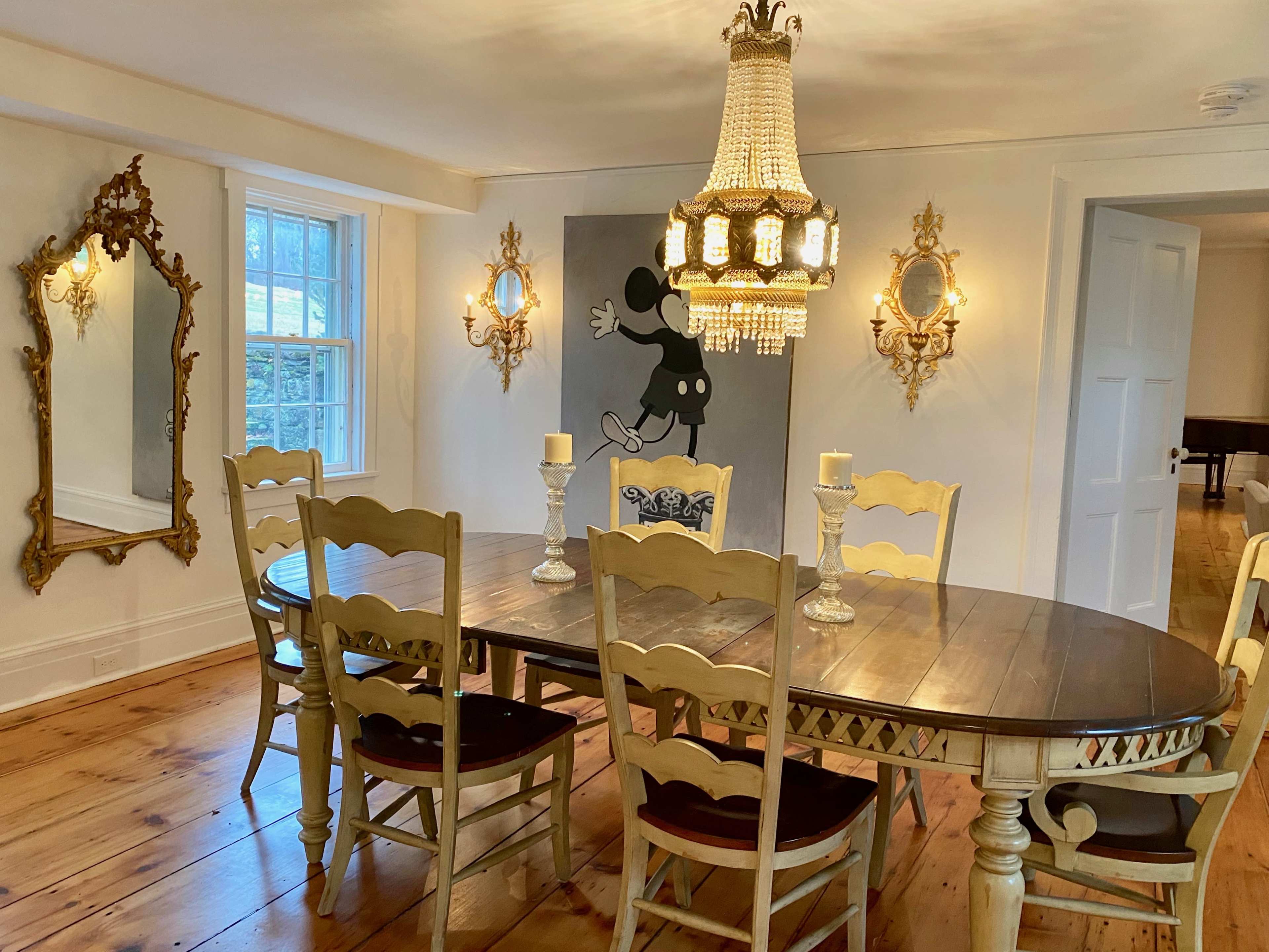 A dining room features a round wooden table surrounded by chairs, with a large chandelier above, mirrors on the walls, and a decorative painting of a cartoon character.