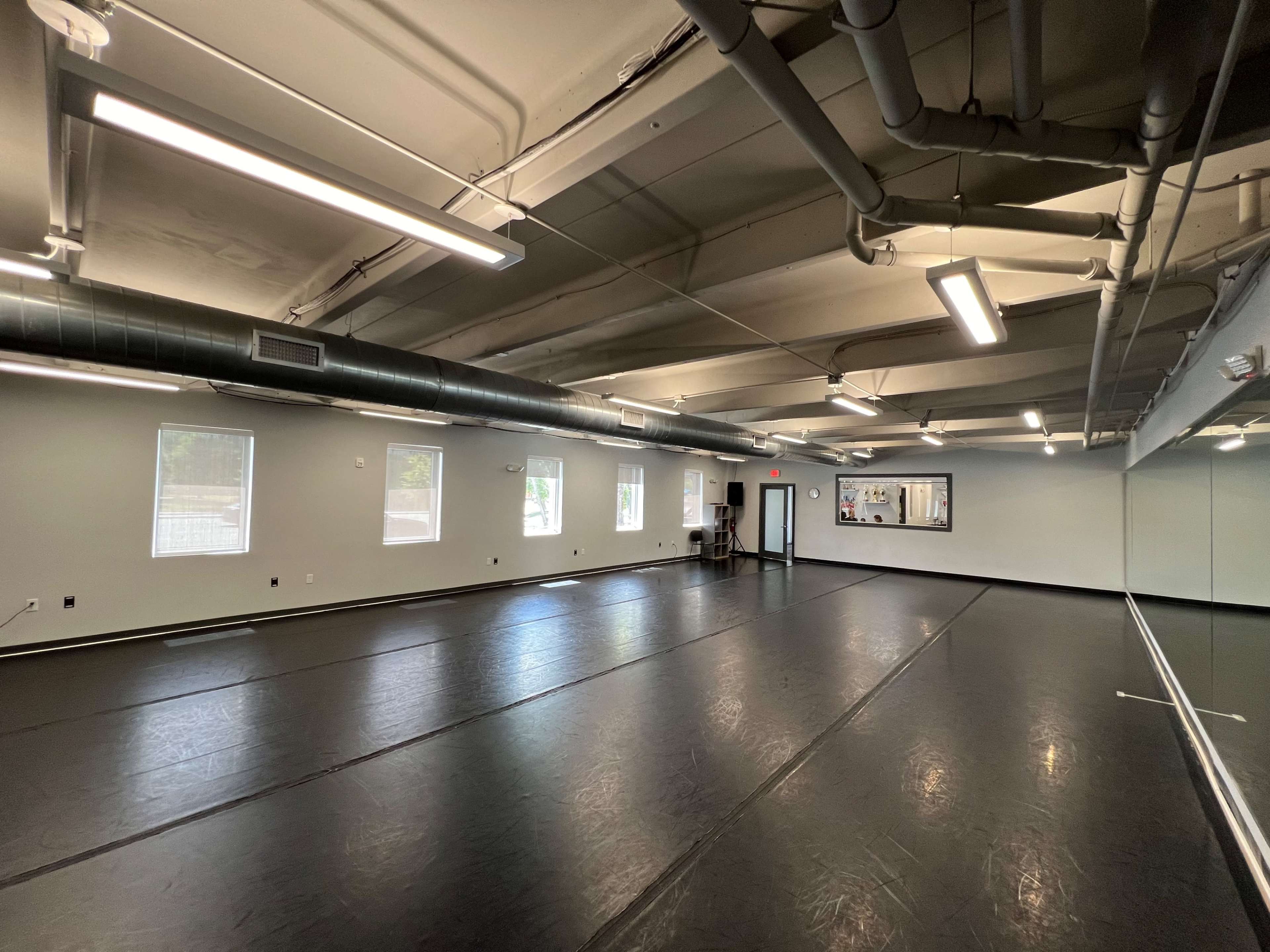 A spacious, empty dance studio with black floors, large mirrors, and windows that allow natural light to enter.