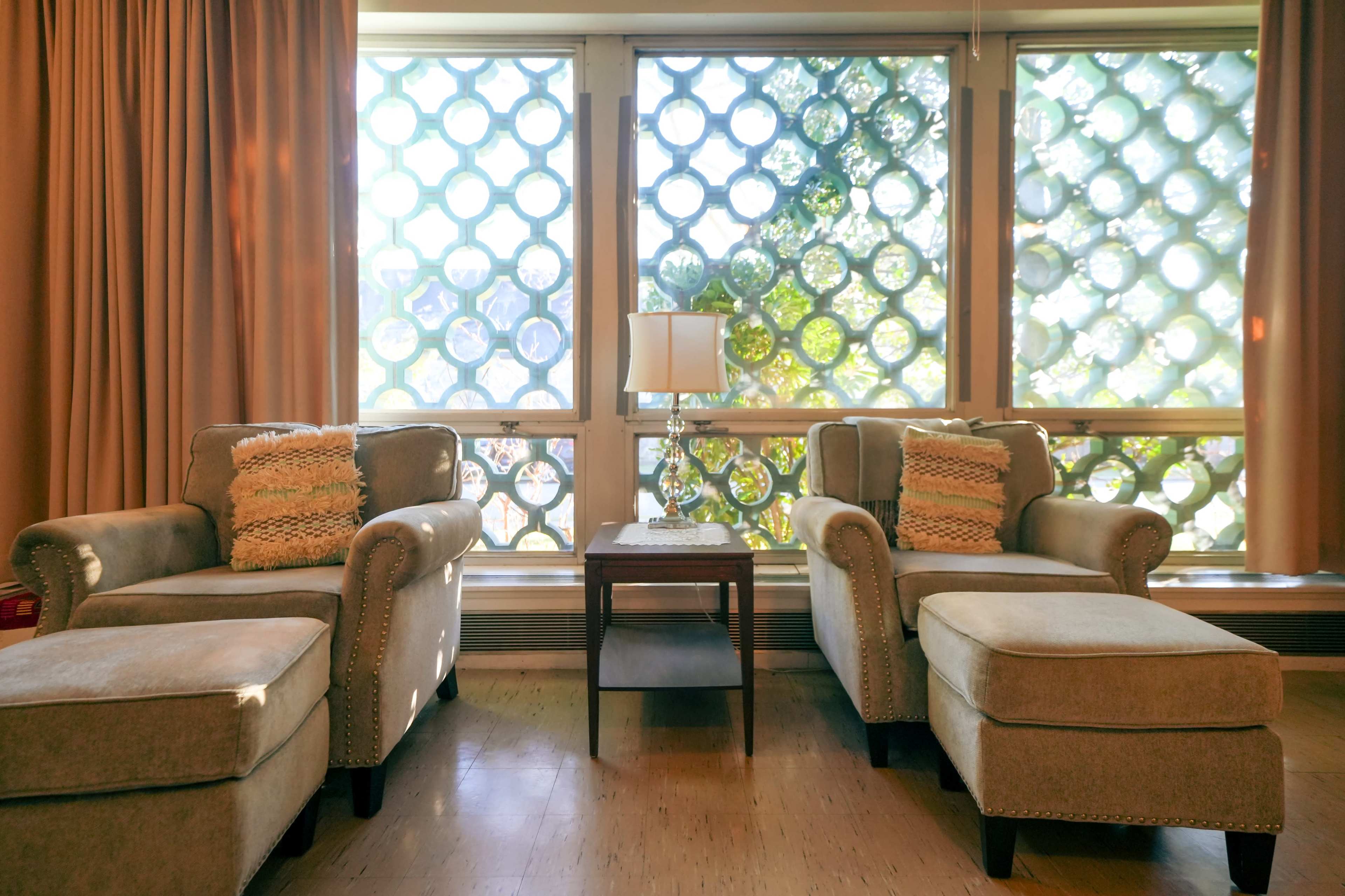 The image shows two upholstered chairs with matching ottomans positioned near large windows featuring a decorative circular lattice design, with a small table and lamp between them.
