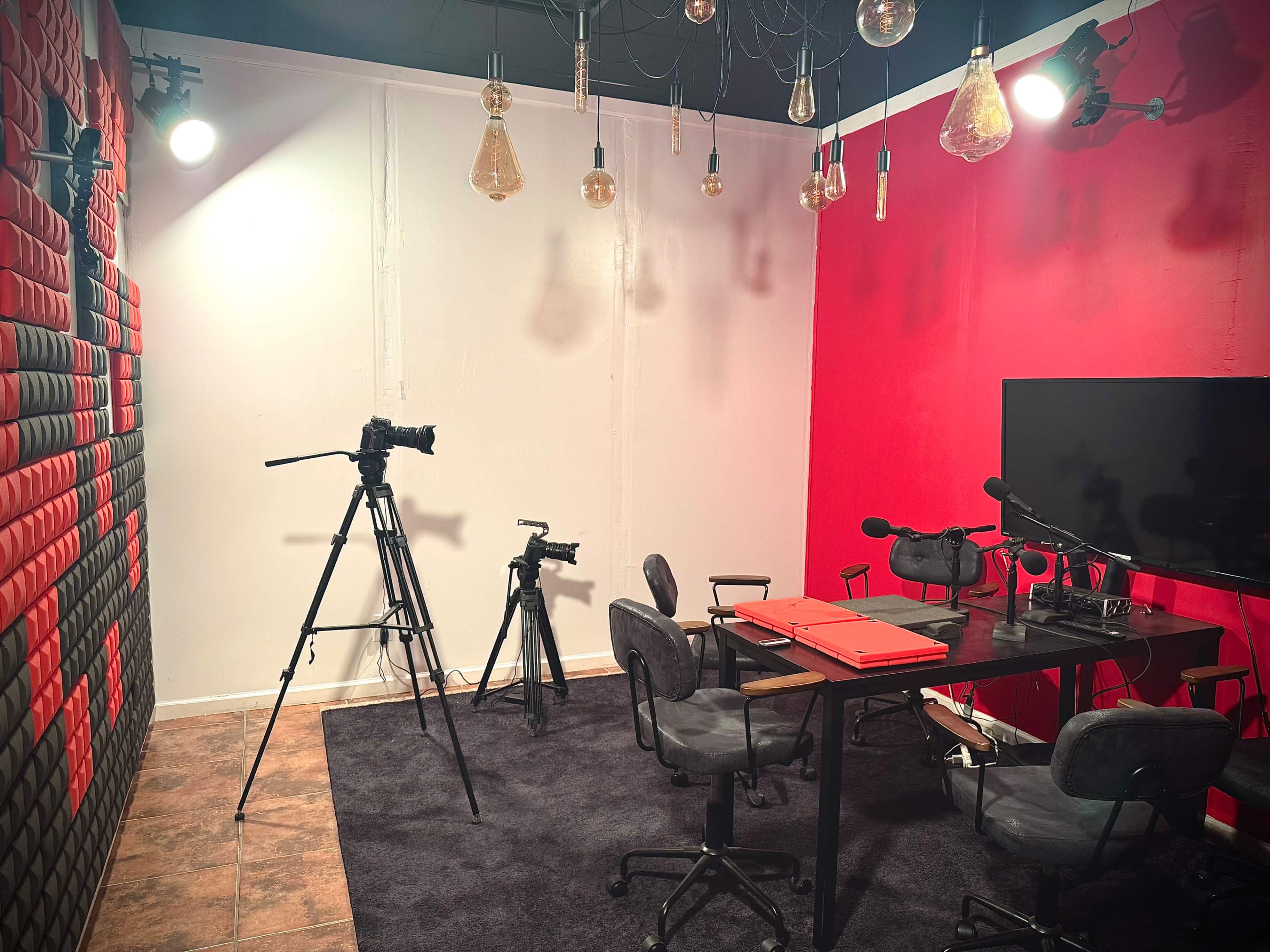 The image shows a media production studio featuring a table with chairs, a camera on a tripod, a TV screen, and pendant lights hanging from the ceiling.