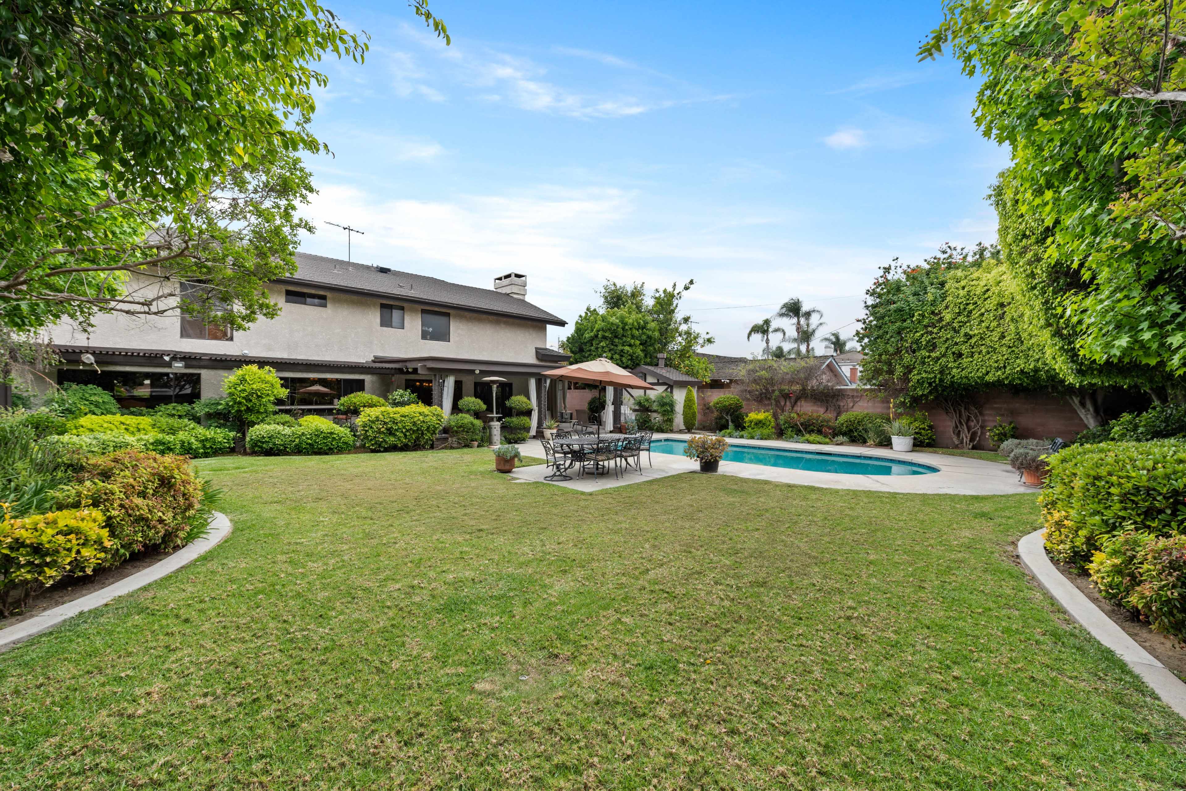The image shows a spacious backyard featuring a manicured lawn, a swimming pool, and a patio area with outdoor furniture.