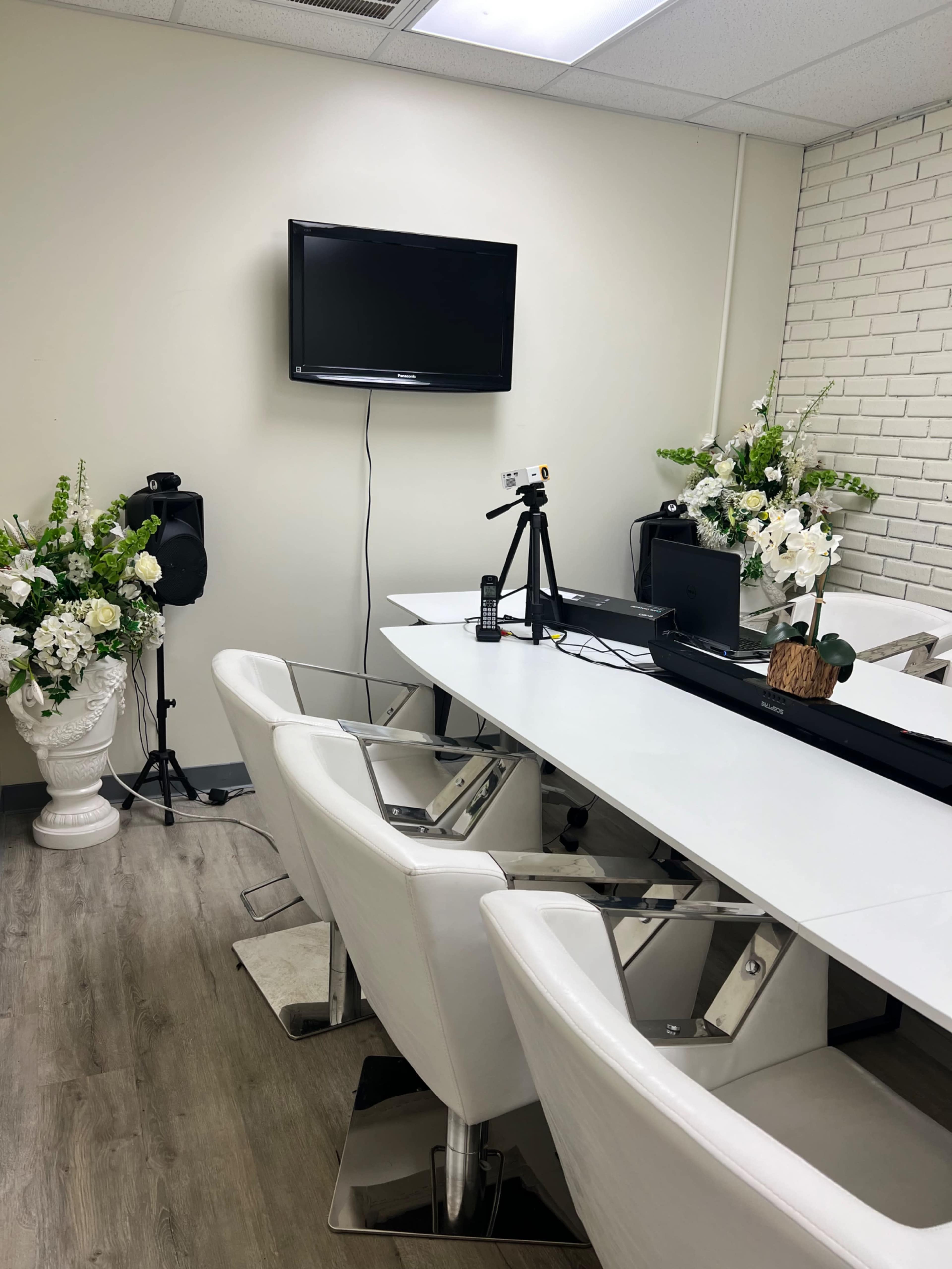 A clean conference room with a long white table, modern chairs, a television mounted on the wall, and decorative flower arrangements on either side.