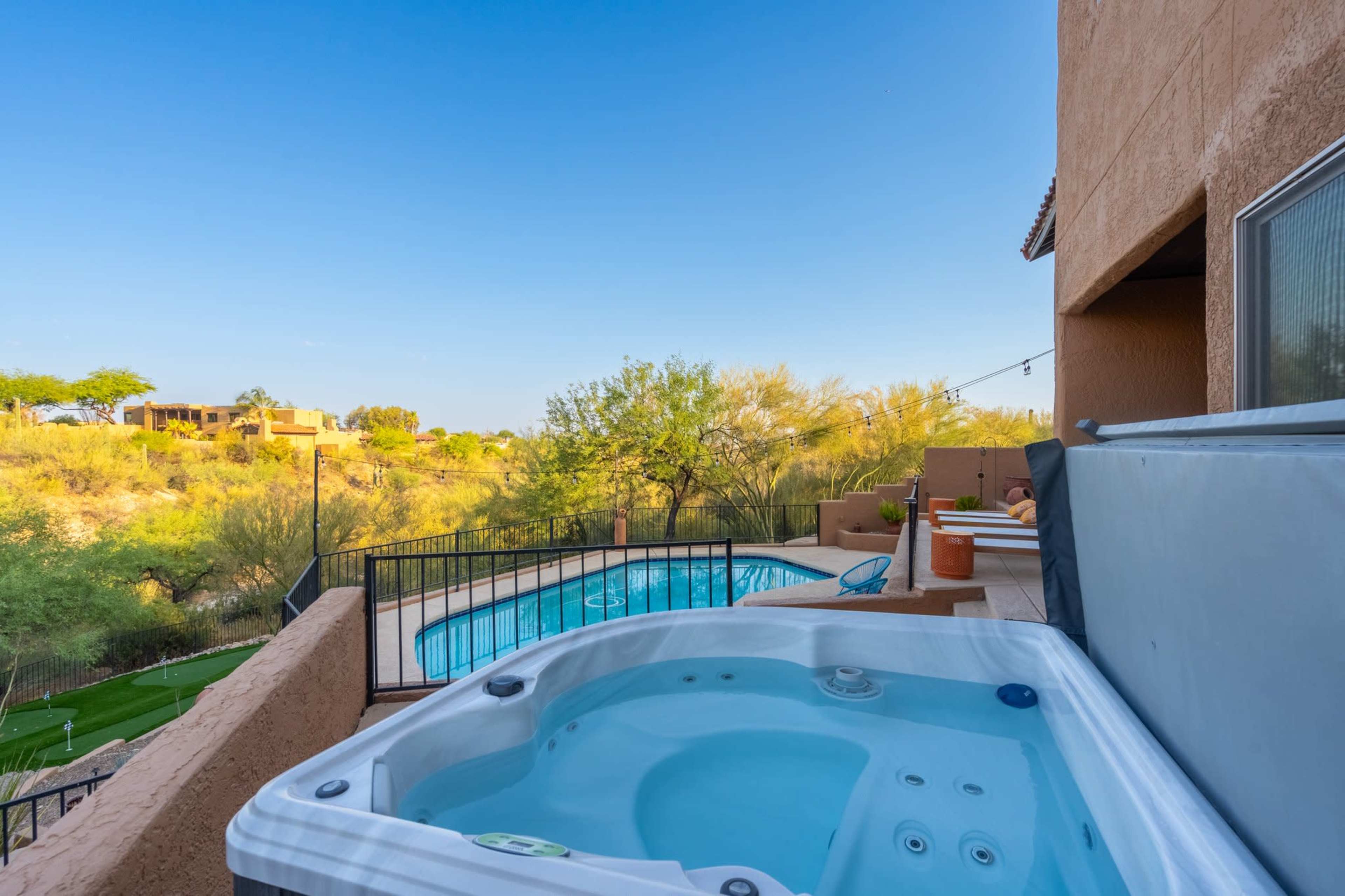 A hot tub sits on a patio overlooking a pool and a desert landscape with greenery in the background.
