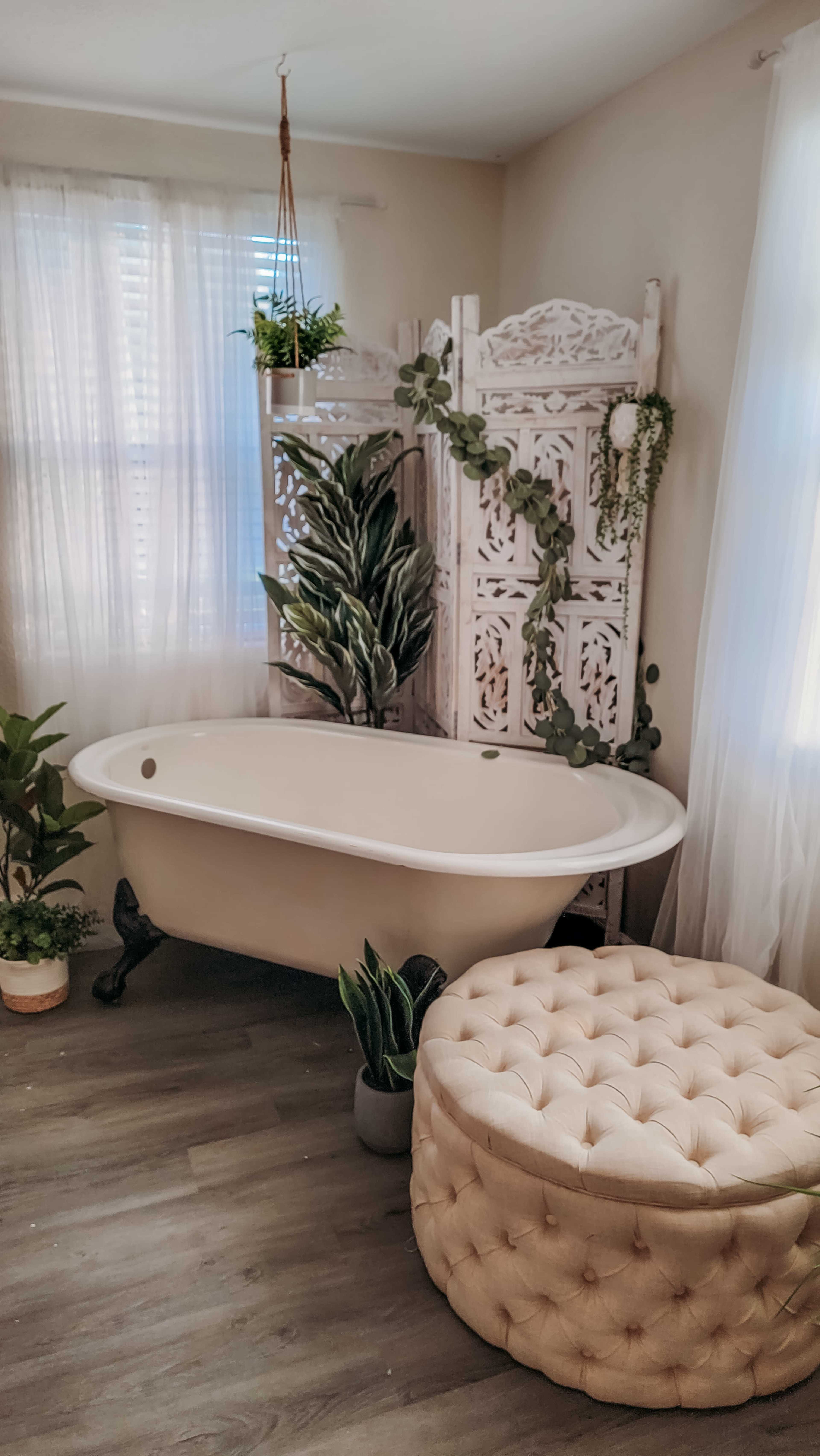 A vintage-style bathtub adjacent to a decorative privacy screen, surrounded by indoor plants and a tufted ottoman in a brightly lit room.