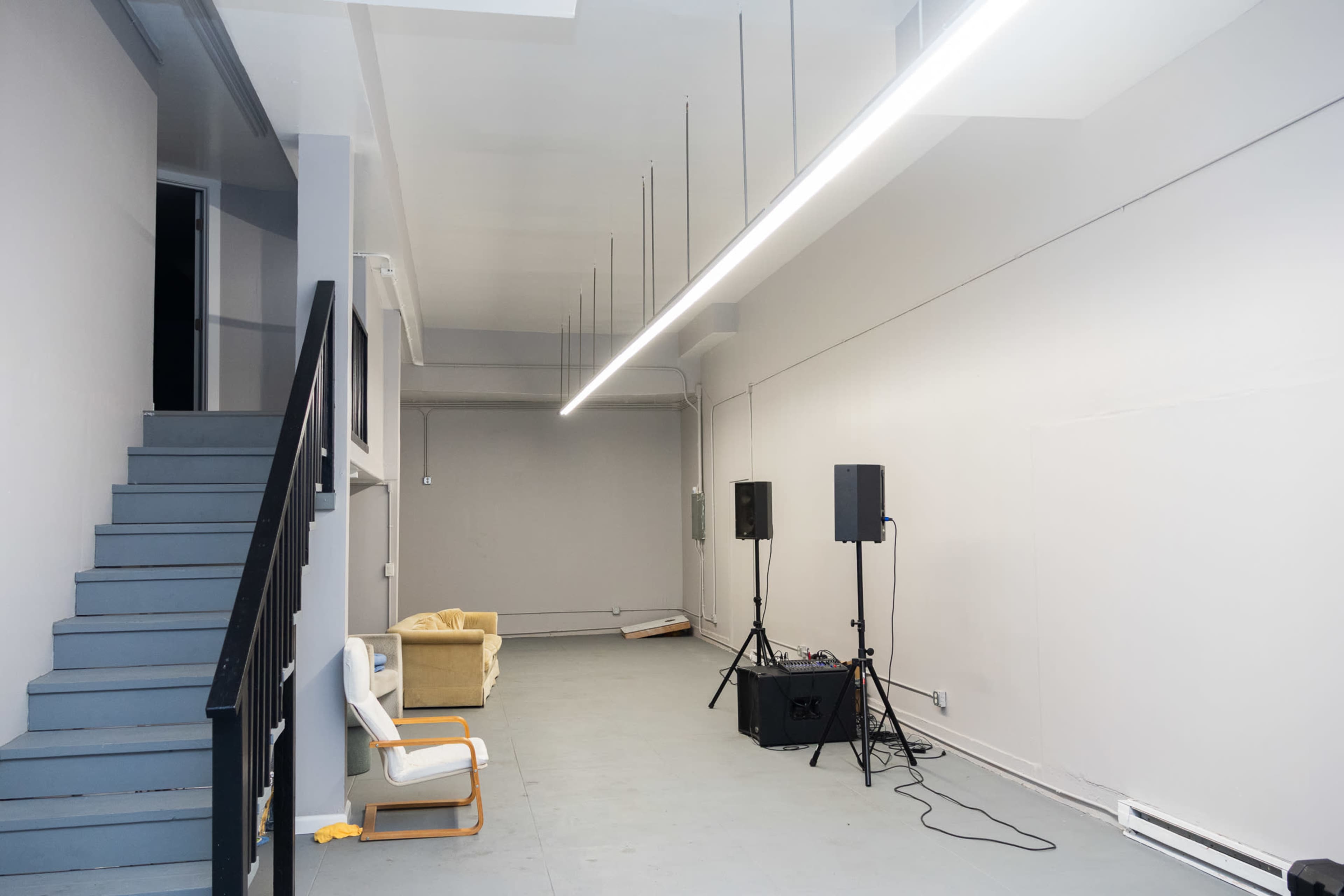 The image shows an empty, well-lit room featuring a staircase, sound equipment, and minimal furnishings.