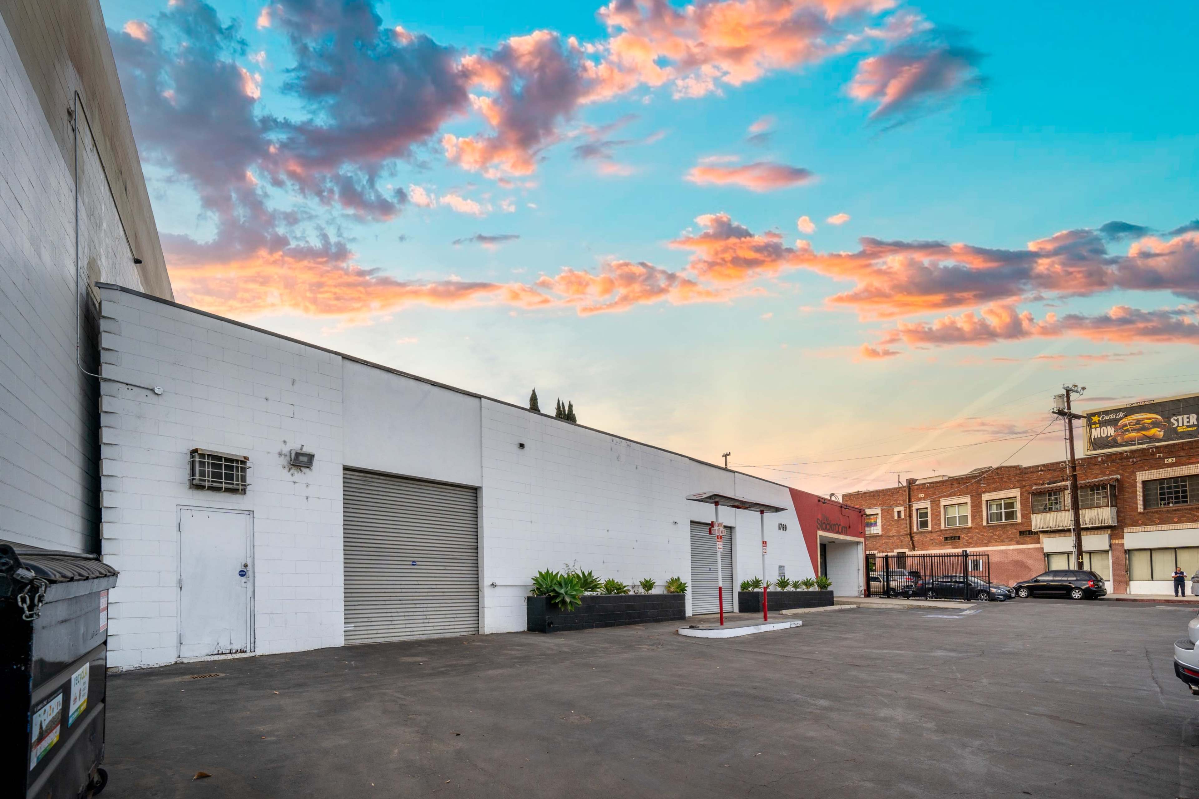 A delivery area with a white wall, a large metal door, a small entrance, potted plants, and a parking lot under a colorful sky at sunset.
