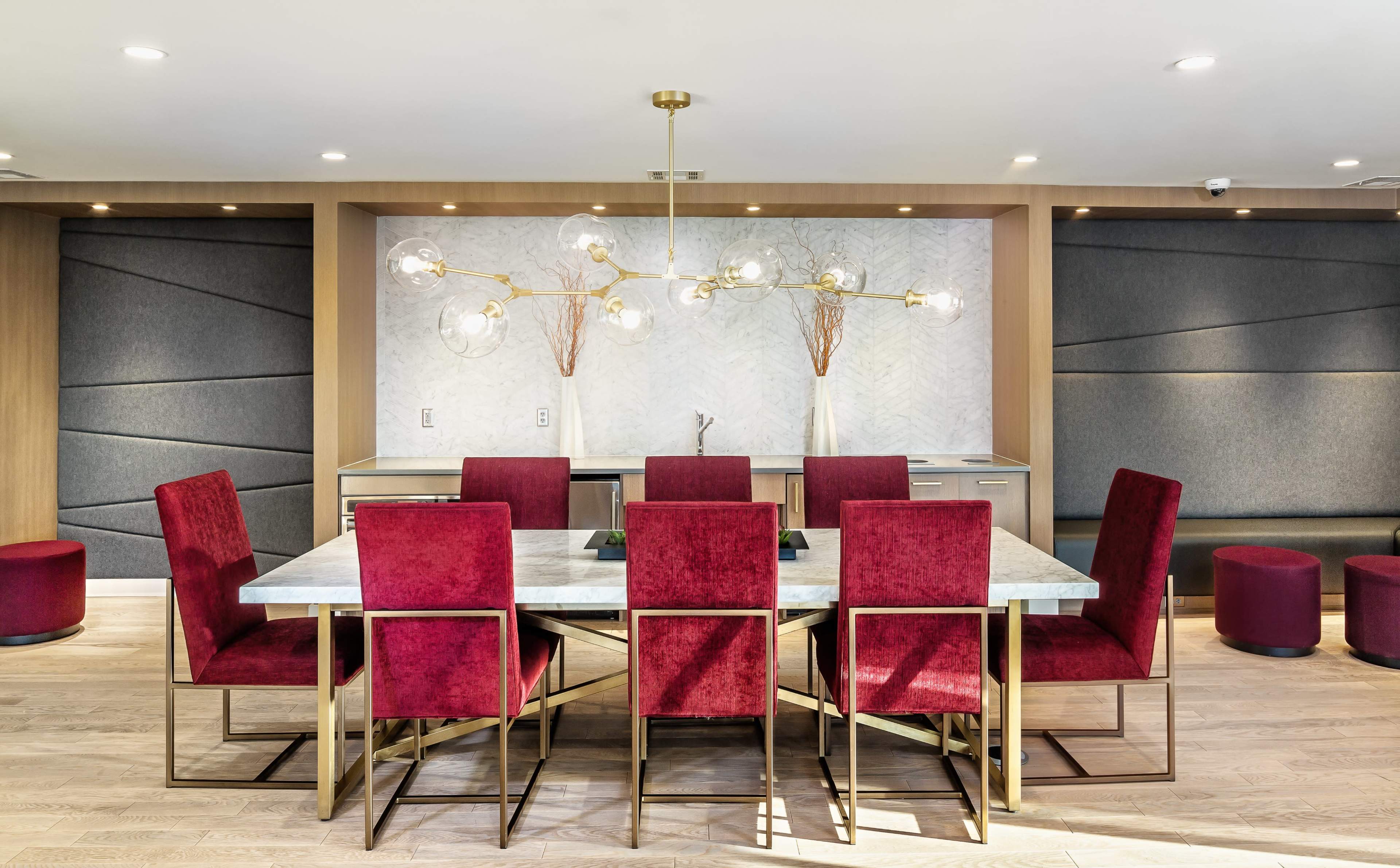 A modern dining area features a marble table surrounded by eight red velvet chairs, with a decorative light fixture hanging above and a textured wall in the background.