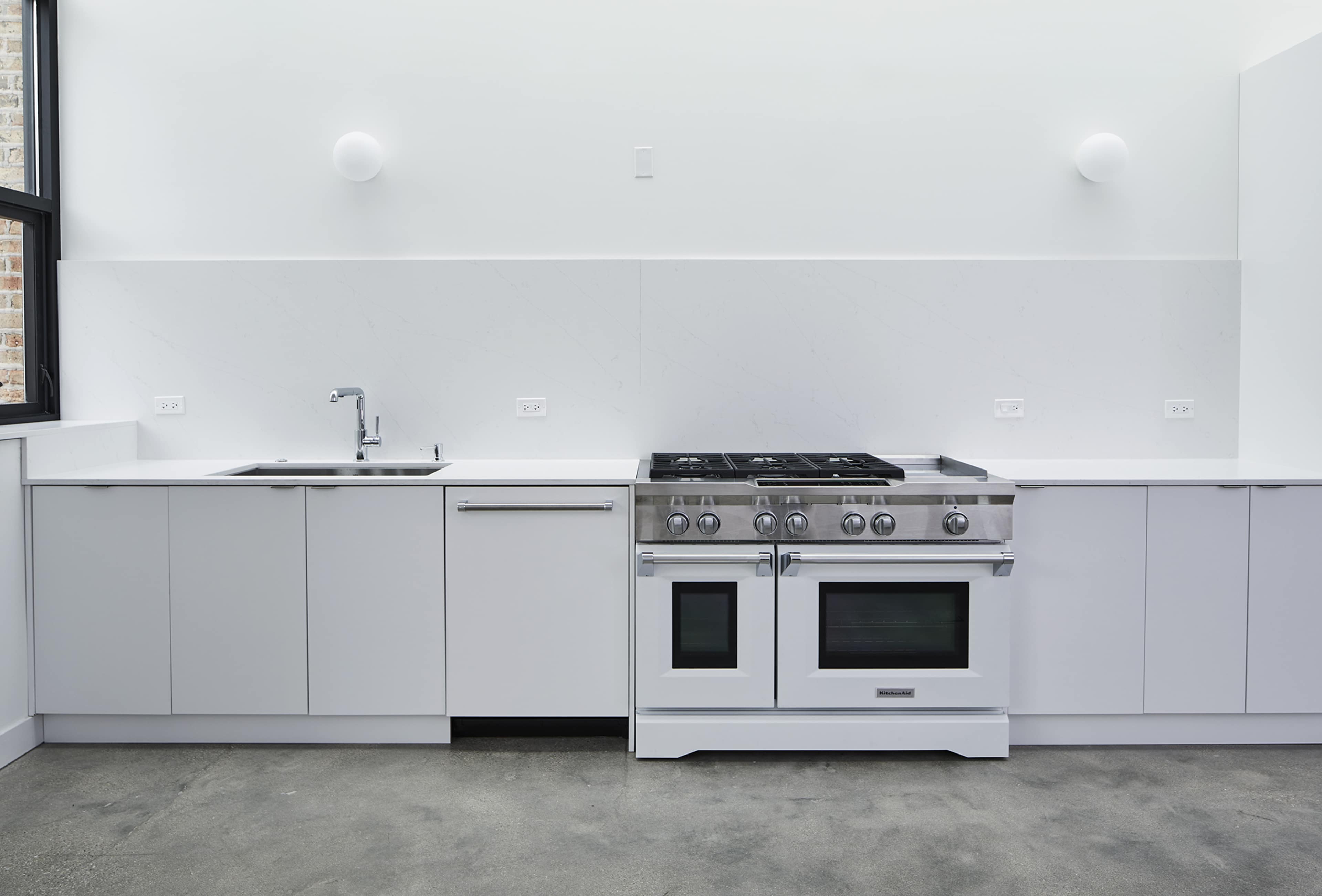 A modern kitchen with white cabinetry, a stainless steel stove, and a single sink under large windows.