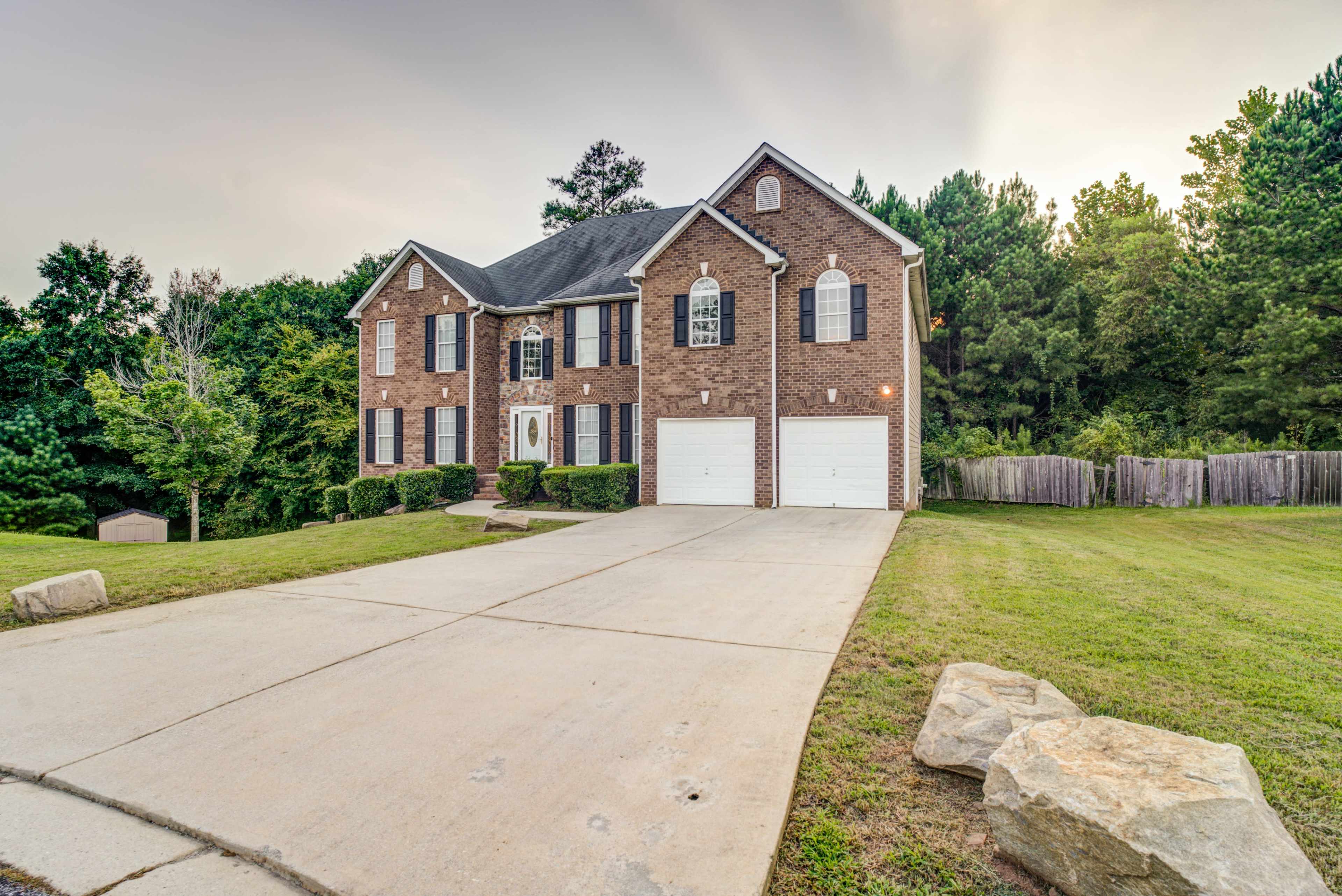 A two-story brick house with black shutters and a concrete driveway is positioned on a grassy lot surrounded by trees.