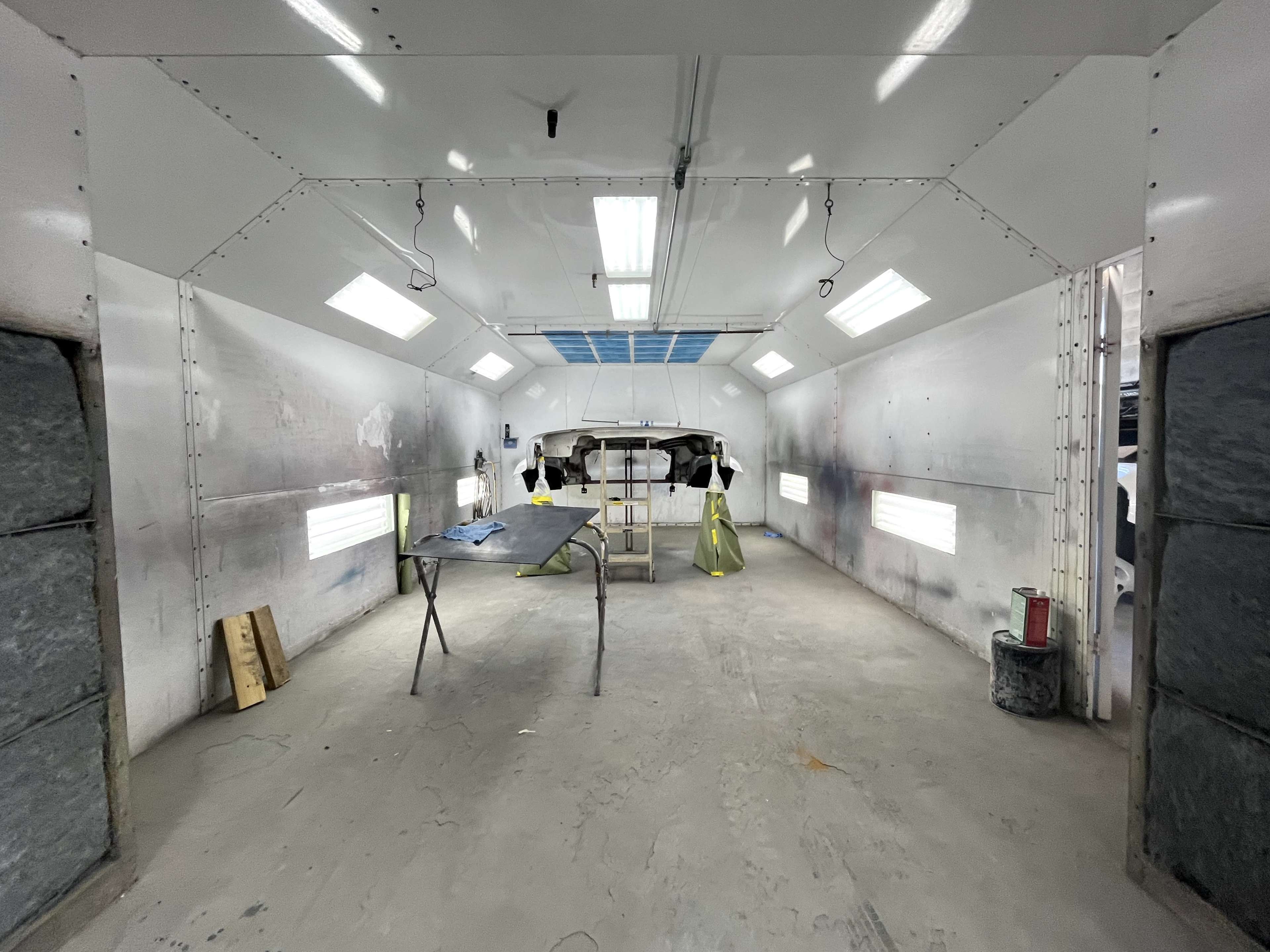 The image shows a spacious automotive paint booth with bright overhead lights and a vehicle partially covered for painting.